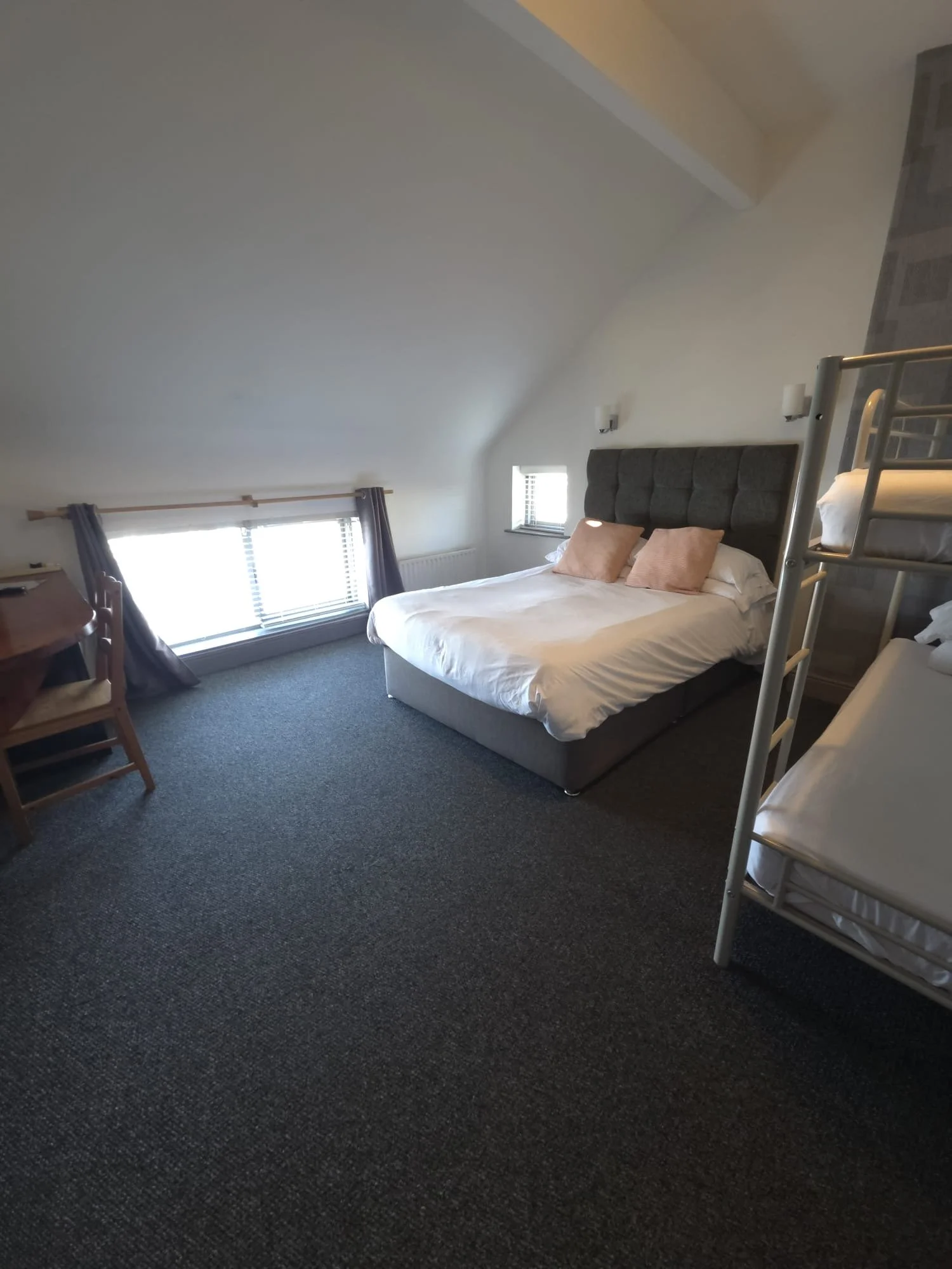 A bedroom with a large bed, pillows, and a headboard, a window with curtains, and bunk beds in the corner, with a carpeted floor and a sloped ceiling.