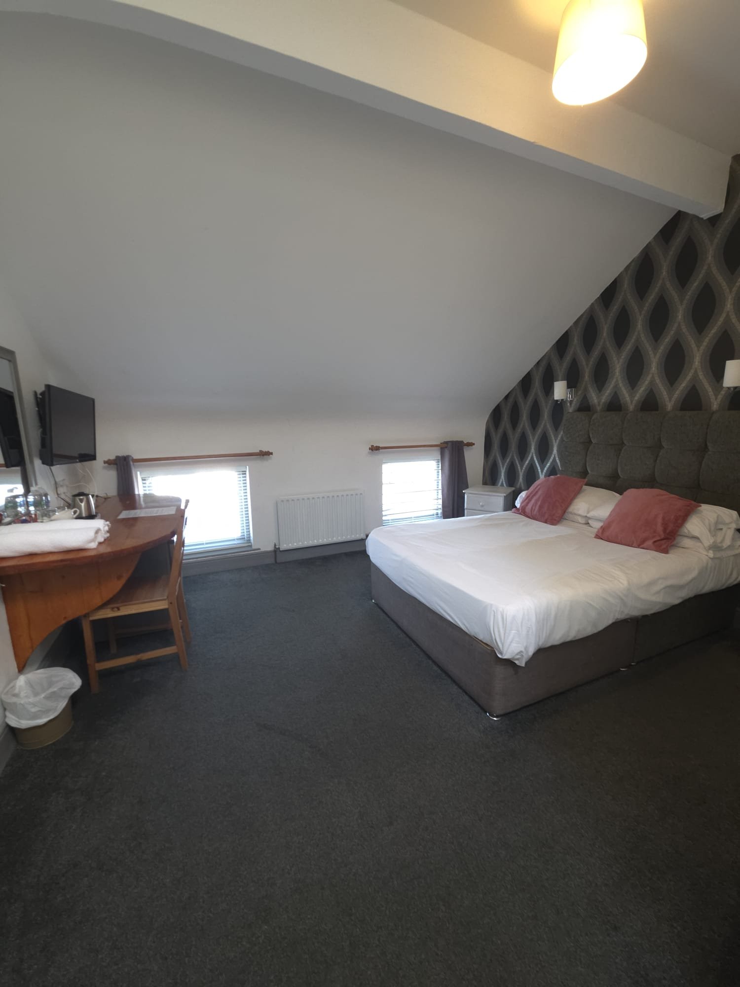 A bedroom with a large bed, nightstand, and a wall-mounted TV, featuring patterned wallpaper and two windows with curtains.