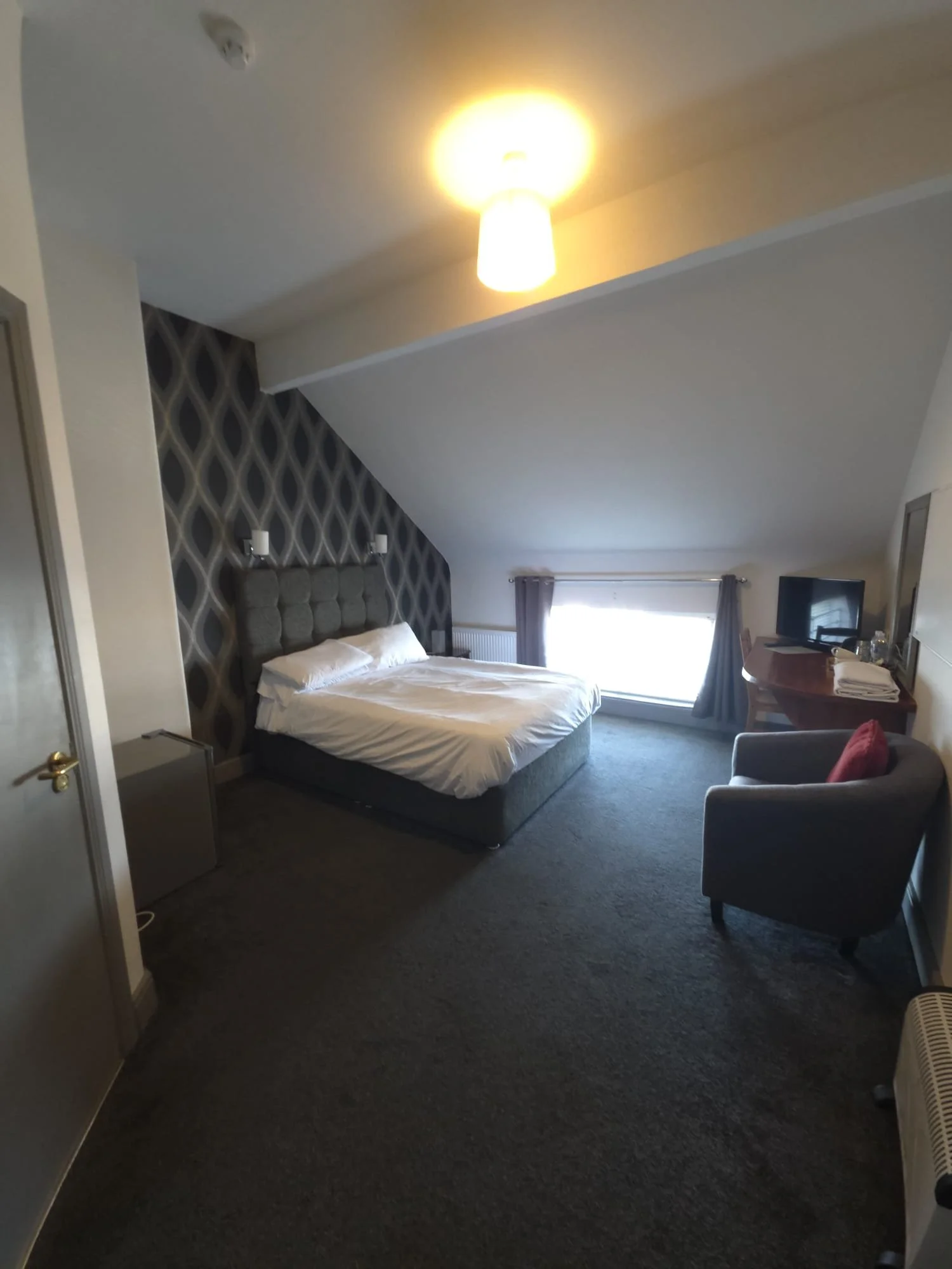 A hotel room with a bed, gray headboard, and white bedding. There is a nightstand next to the bed with a lamp. The room has a window with curtains and a small TV on a wooden desk. A gray armchair with a red cushion is near the window.