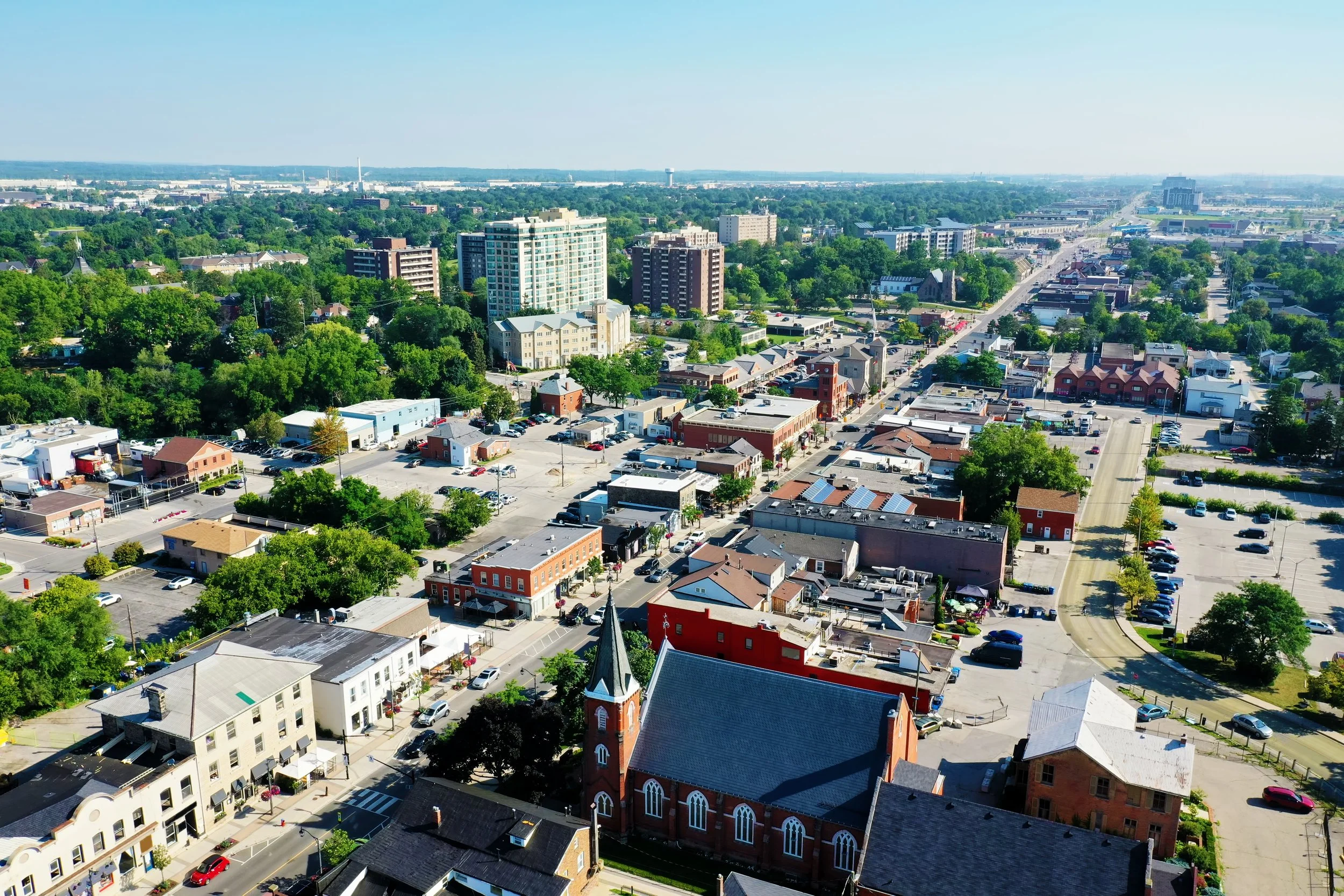 Aerial cityscape view showing a small downtown area with a prominent red brick church and surrounding buildings, parking lots, and tree-lined streets, extending to tall modern buildings and greenery in the background under a clear blue sky.