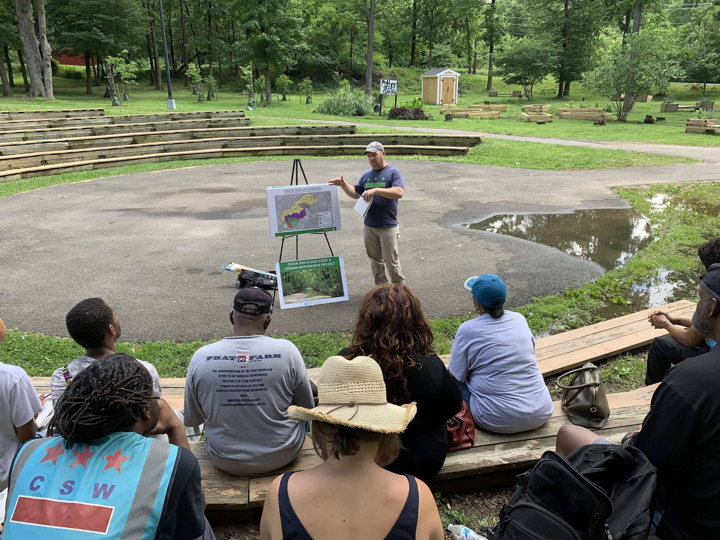 A man giving a presentation outdoors to a small audience sitting on wooden benches near a pond and surrounded by trees, with maps and informational posters displayed on an easel.