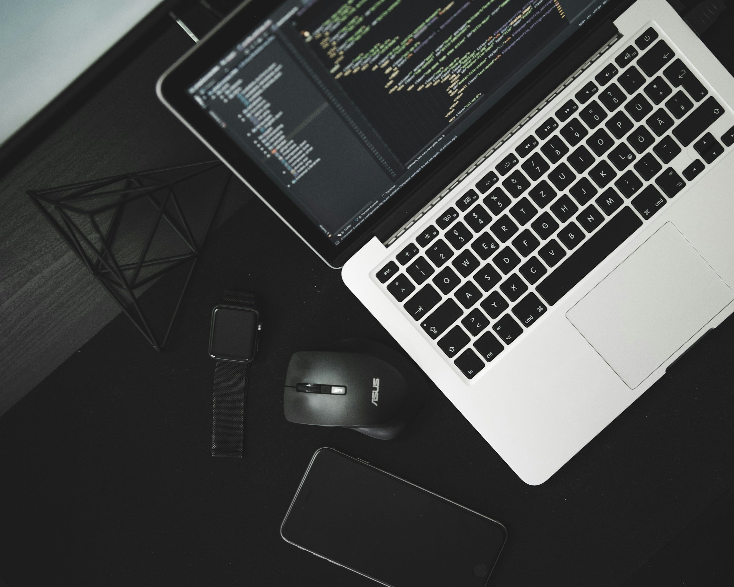 A laptop computer with a code editor open on the screen, placed on a black desk next to a geometric metal wire holder.