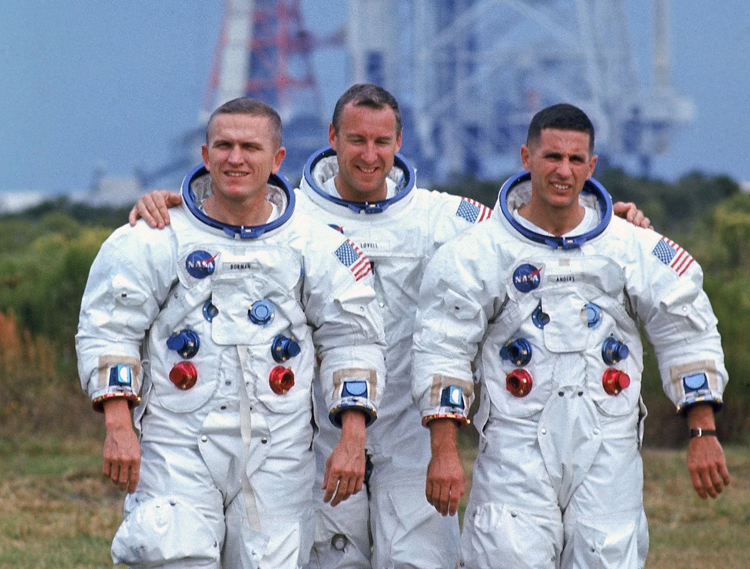 Three astronauts in white space suits standing outdoors with their arms around each other, smiling.