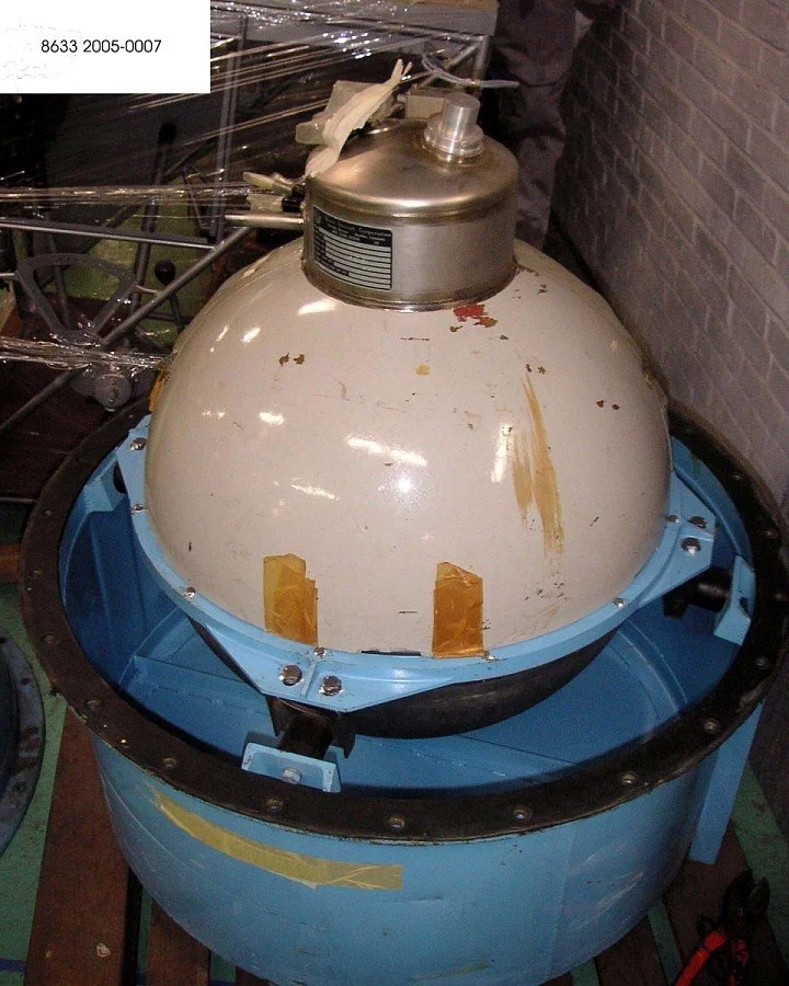 A large spherical spacecraft component with a metallic top mounted on a blue support structure, secured with tape and bolts, in a workshop or lab environment.