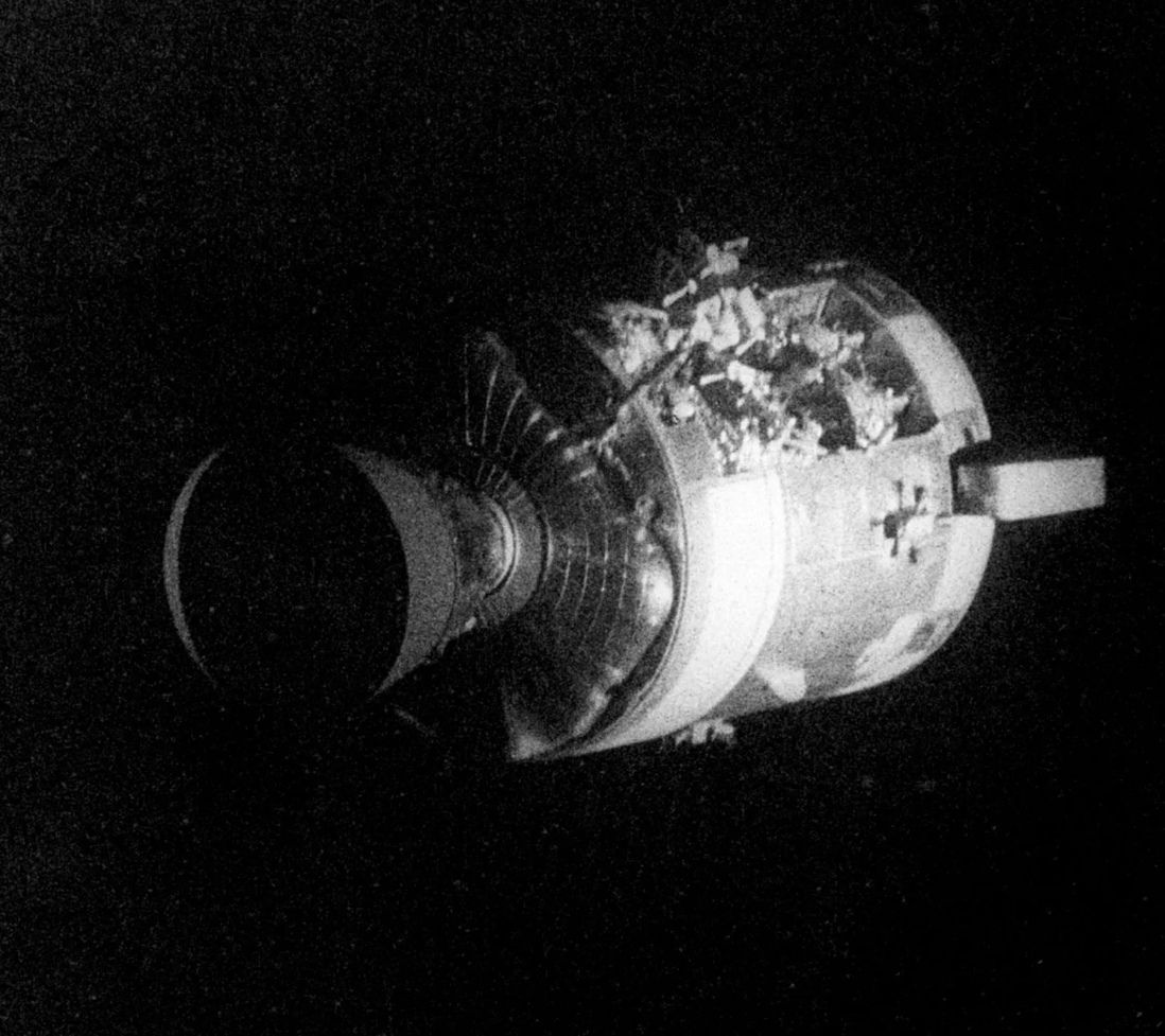 A black and white photograph of a spacecraft or space capsule with astronauts inspecting it in space.