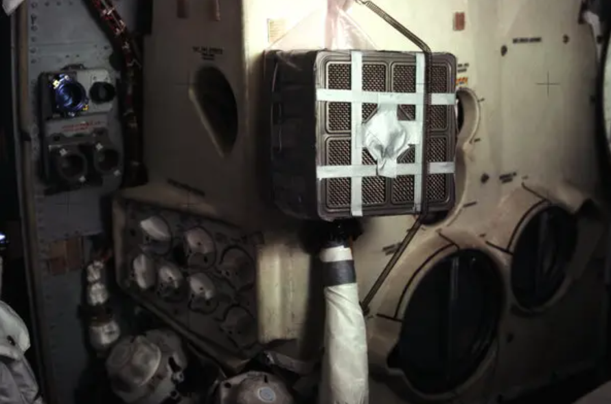 View inside the International Space Station showing a metal wall with circular vents, storage containers, and a small black box with white tape.