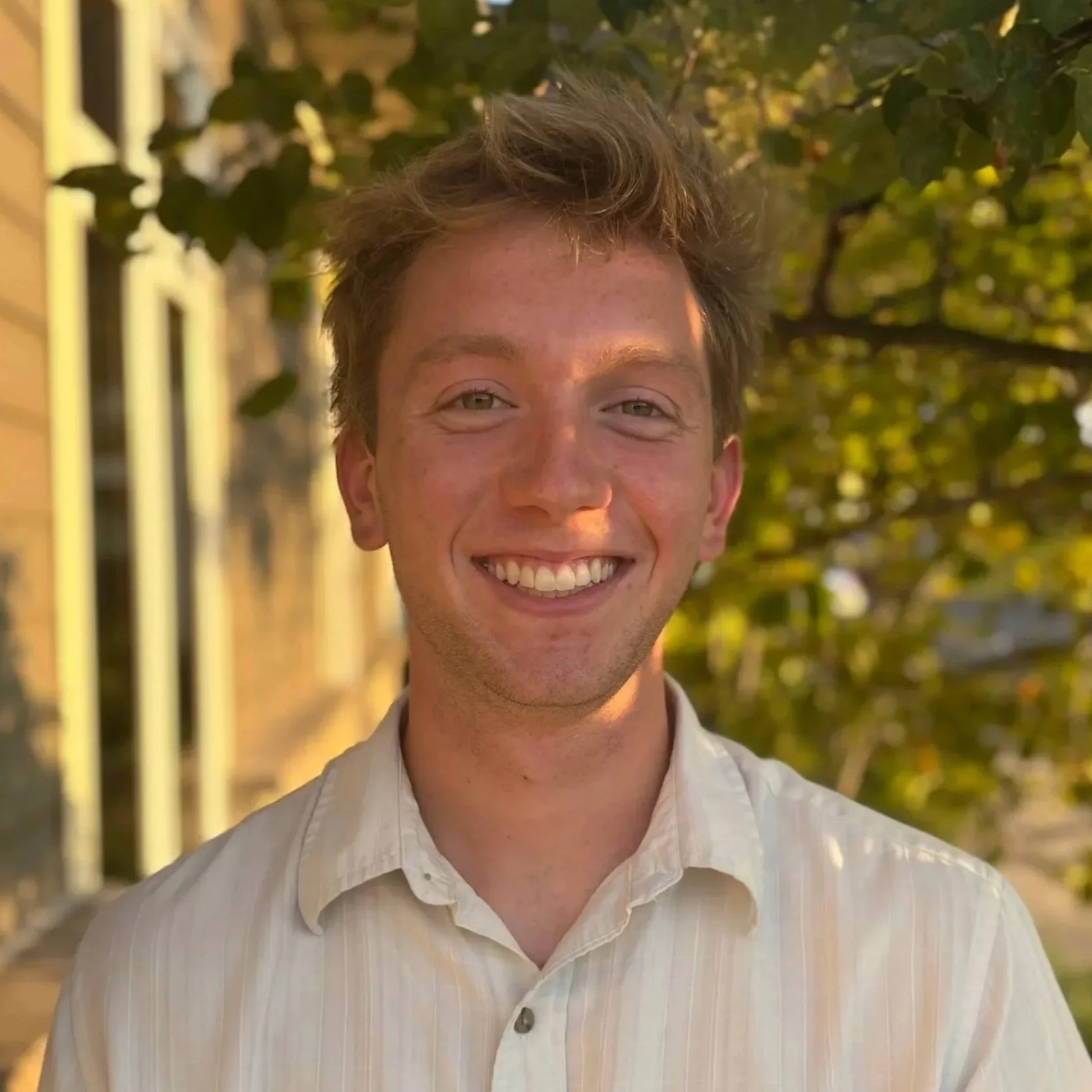 A smiling man with short brown hair and a beard wearing a blue plaid shirt standing outside near a house with trees in the background.