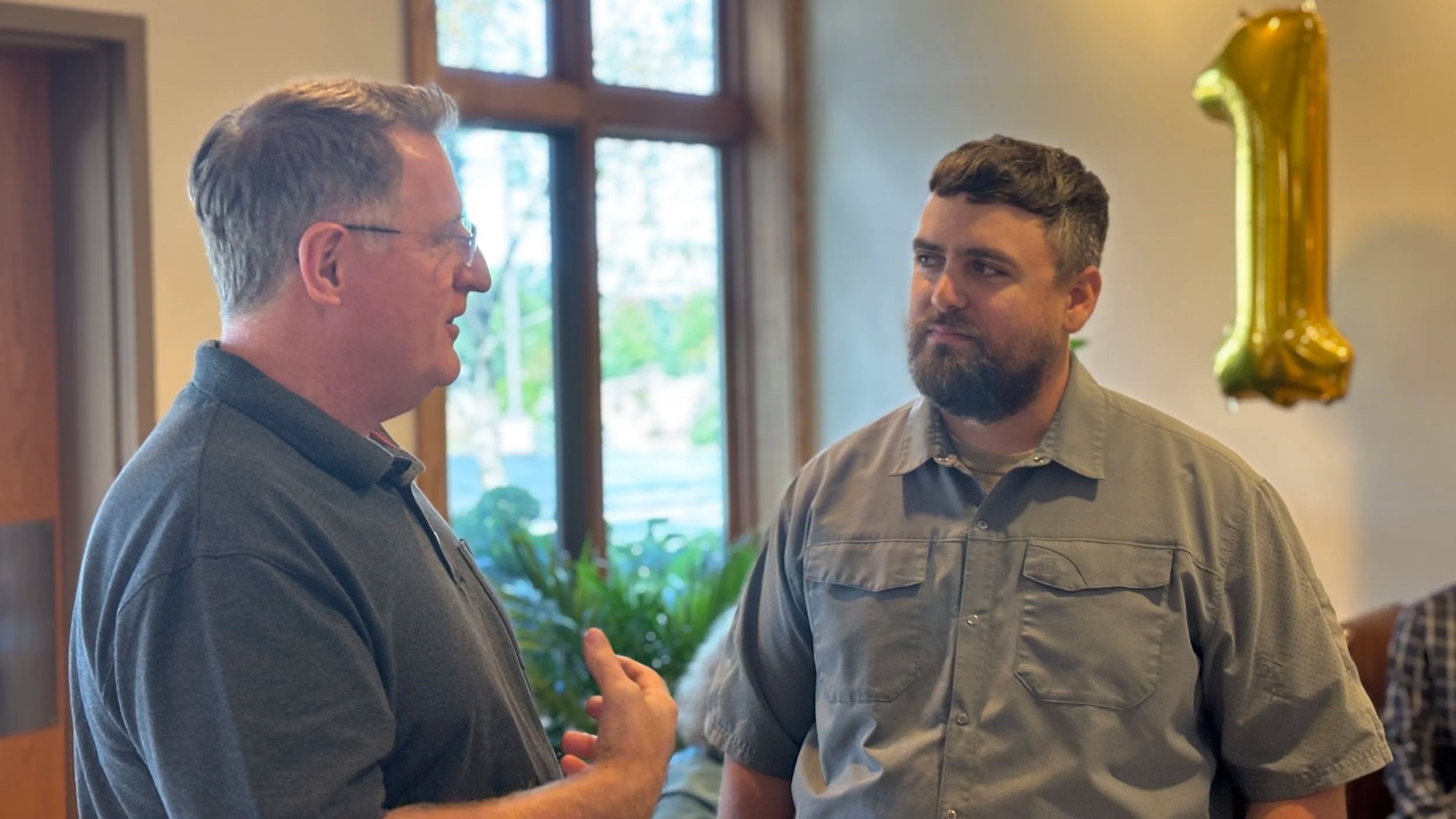 Two men are conversing indoors at a celebration. One man is wearing a dark gray polo shirt, glasses, and has short hair. The other man has a beard, short dark hair, and is wearing a gray button-up shirt. A large gold balloon shaped as the number one is hanging in the background.
