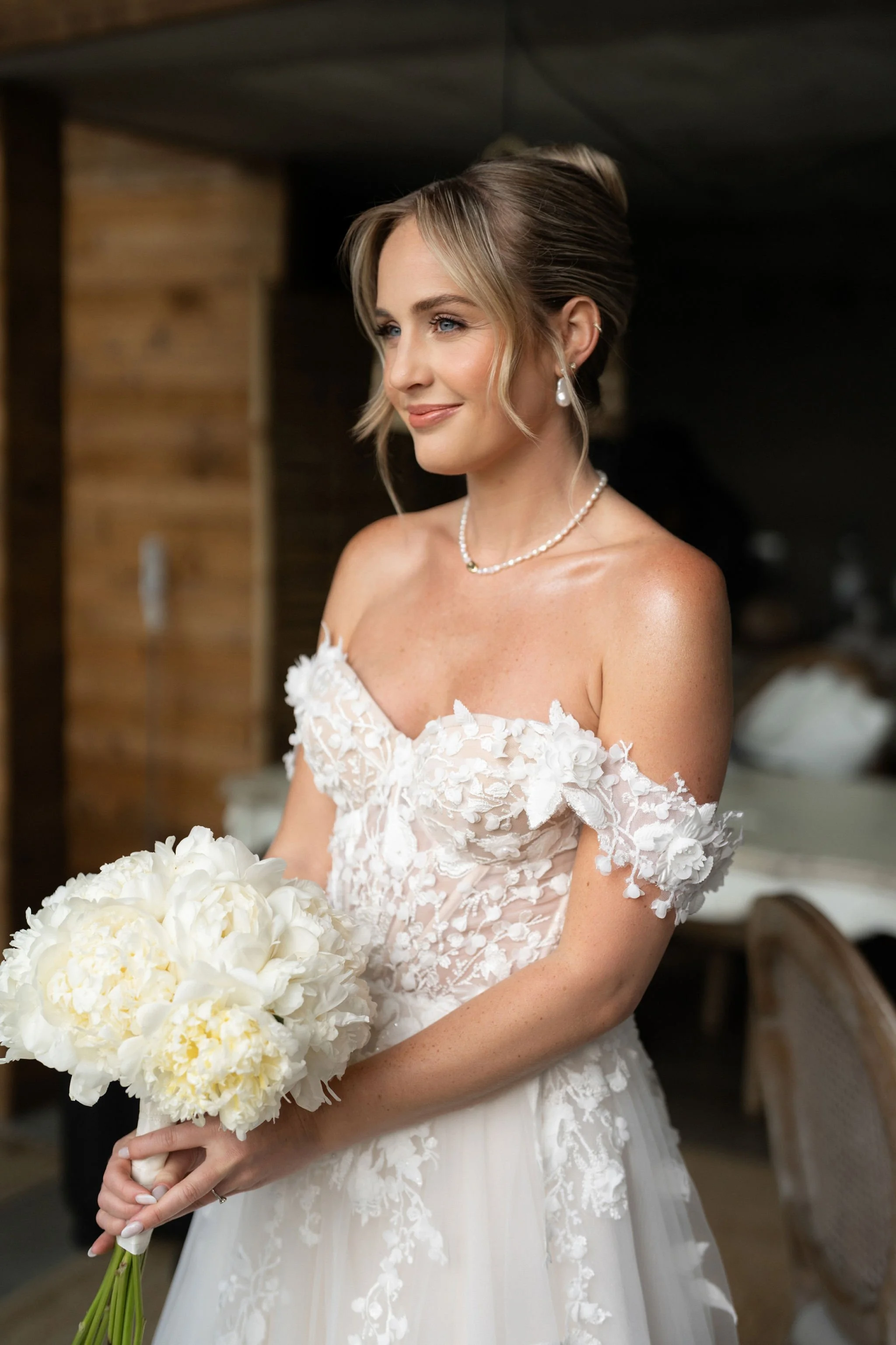 A bride in an off-the-shoulder white wedding dress holding a bouquet of white flowers, smiling softly.