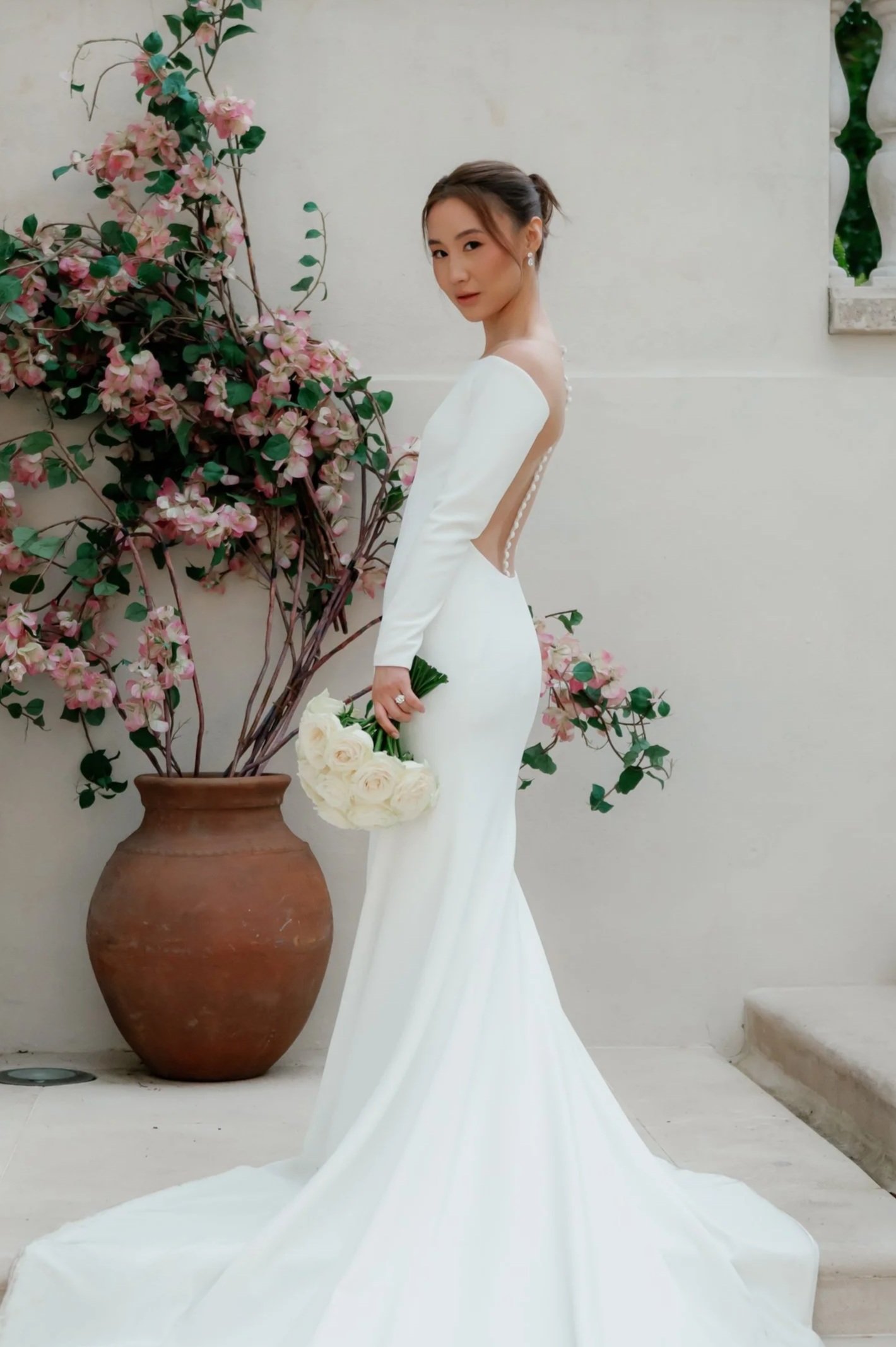 A bride in a white wedding dress holding a bouquet of white roses, standing near a large clay pot with pink flowers and greenery, against a plain wall.