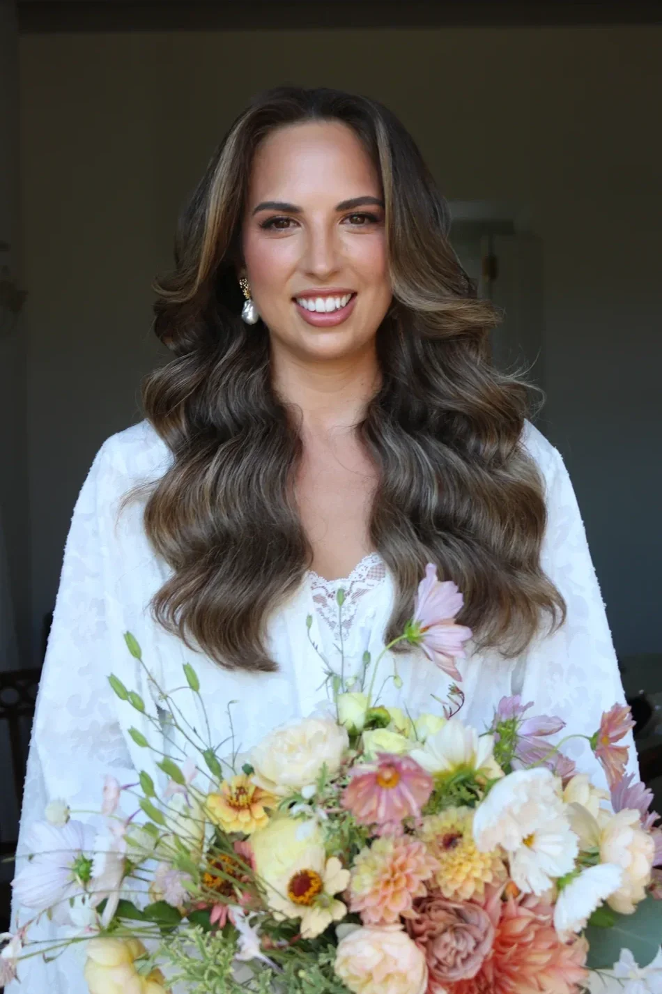 A woman with long wavy brown hair, wearing pearl earrings and a white lace top, holding a bouquet of pink, cream, and peach flowers, smiling at the camera.
