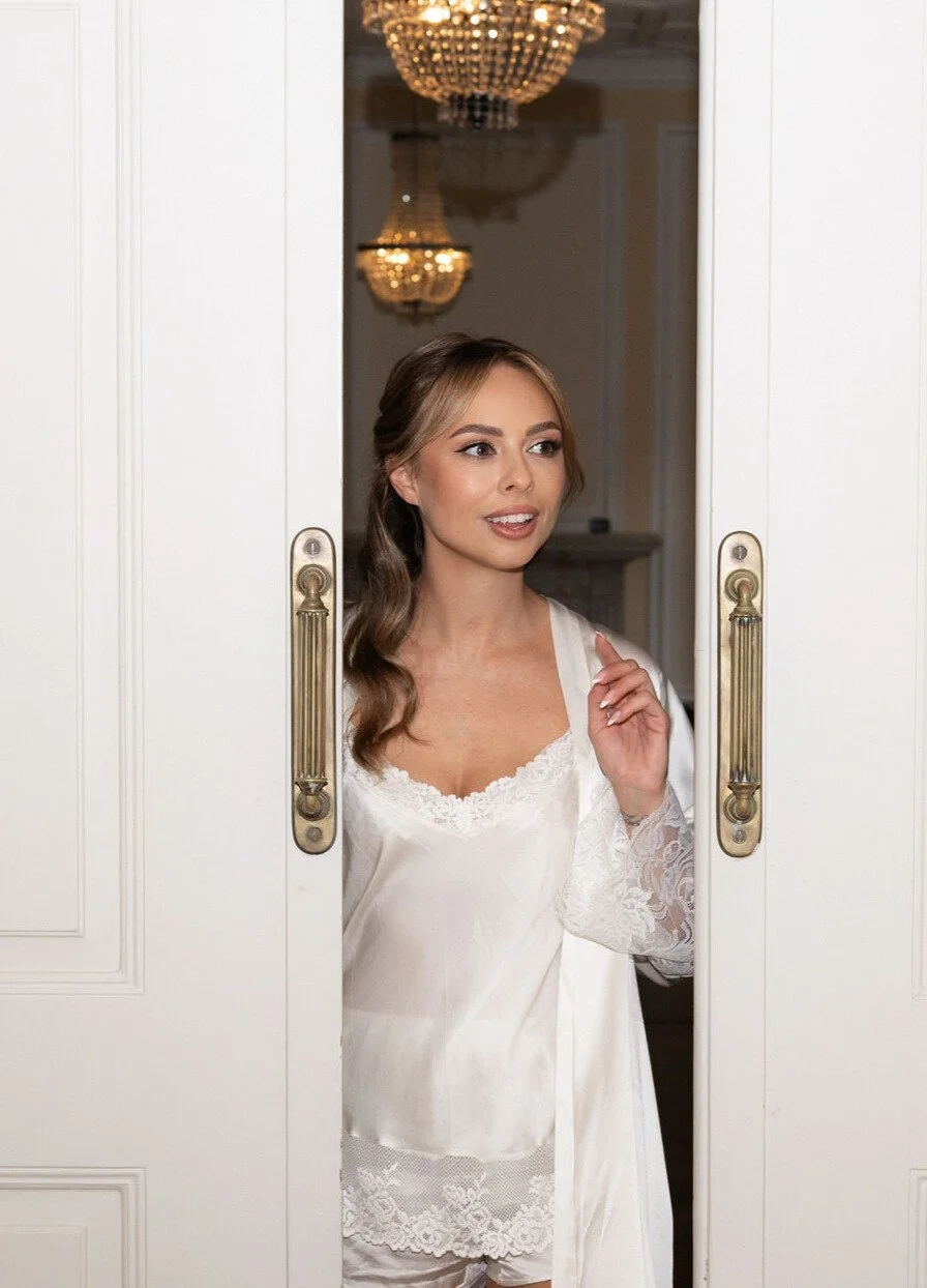 A woman with long wavy brown hair and makeup stands in a doorway, wearing a white lace-trimmed satin nightgown, looking to her right with a slight smile.