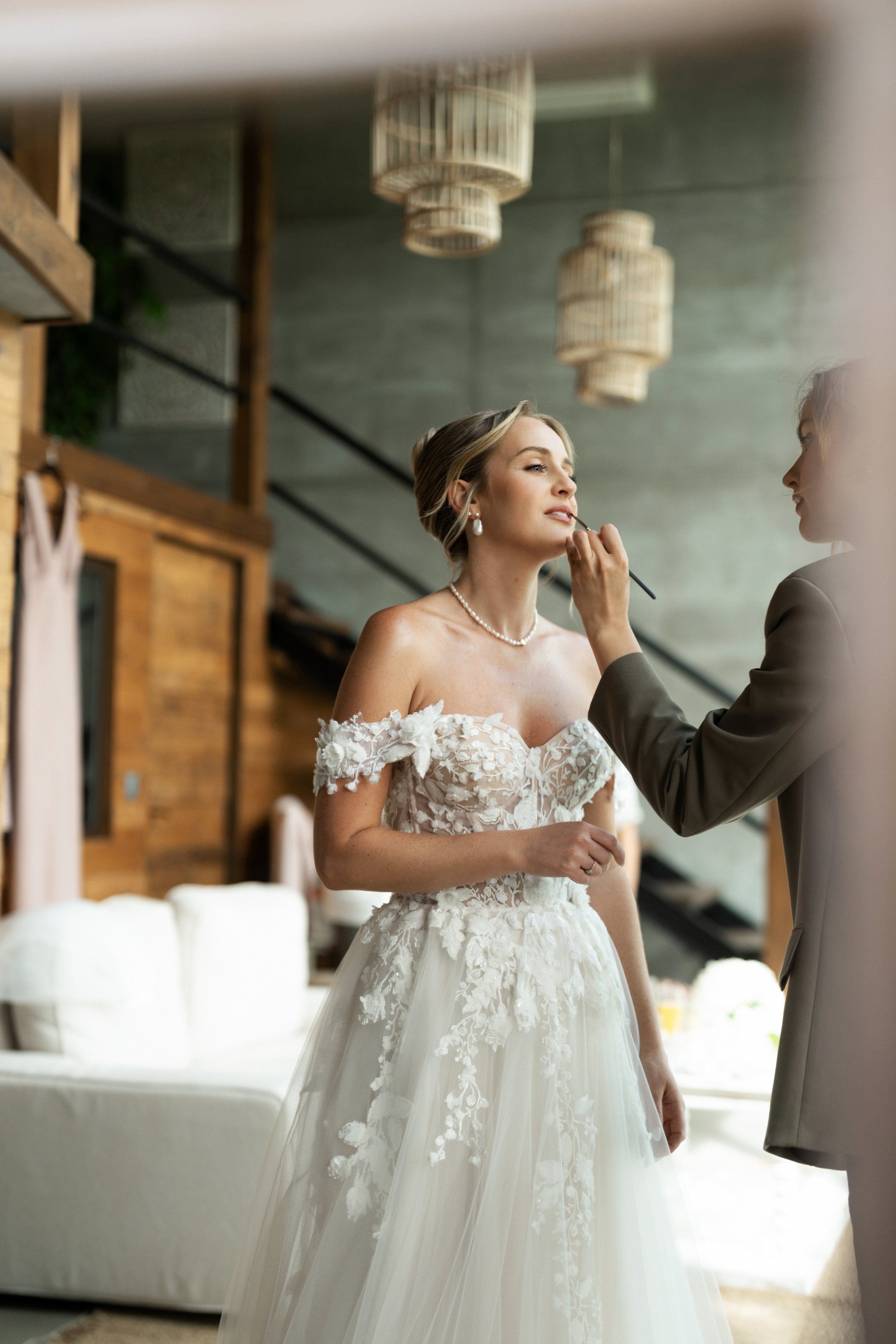 A bride wearing a white wedding dress with floral lace details is having makeup applied by a makeup artist in a modern, rustic-style room.