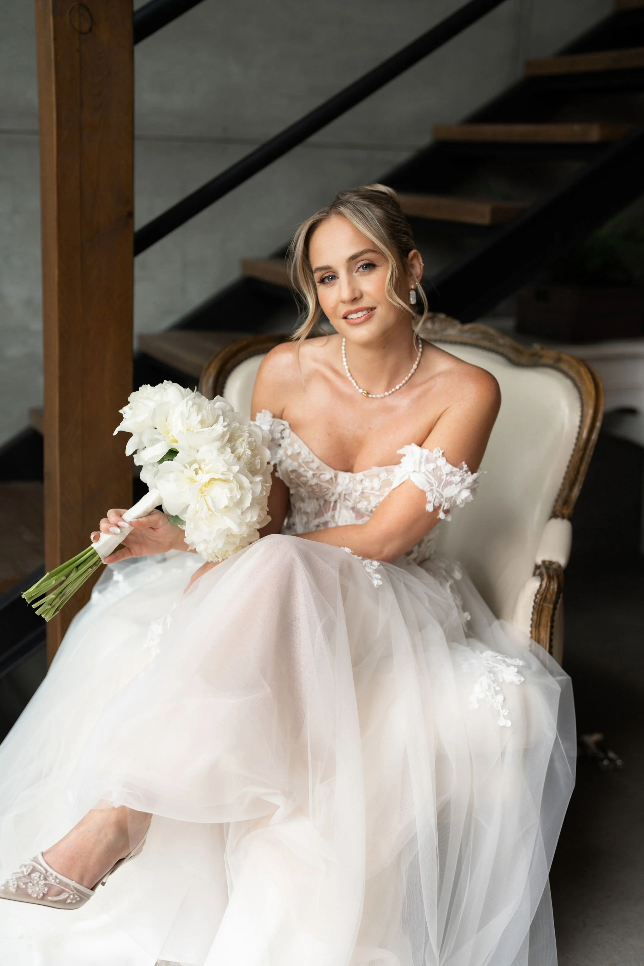 A bride sitting on an elegant vintage armchair, wearing a white wedding dress with lace details, holding a bouquet of white flowers, smiling softly, indoors with a staircase in the background.