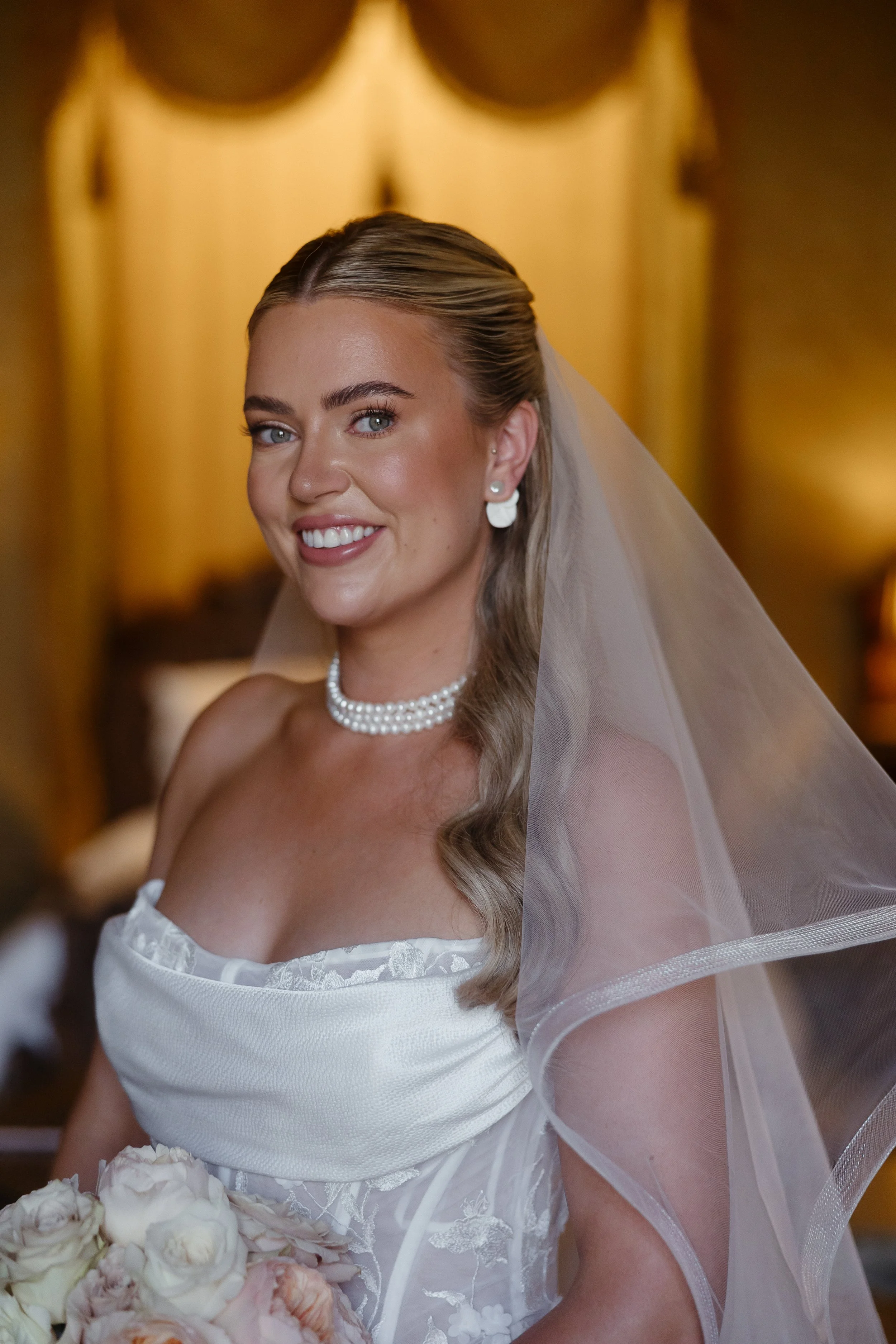 A smiling bride in a white wedding dress, wearing pearl earrings and necklace, with a veil, holding a bouquet of white and blush roses.