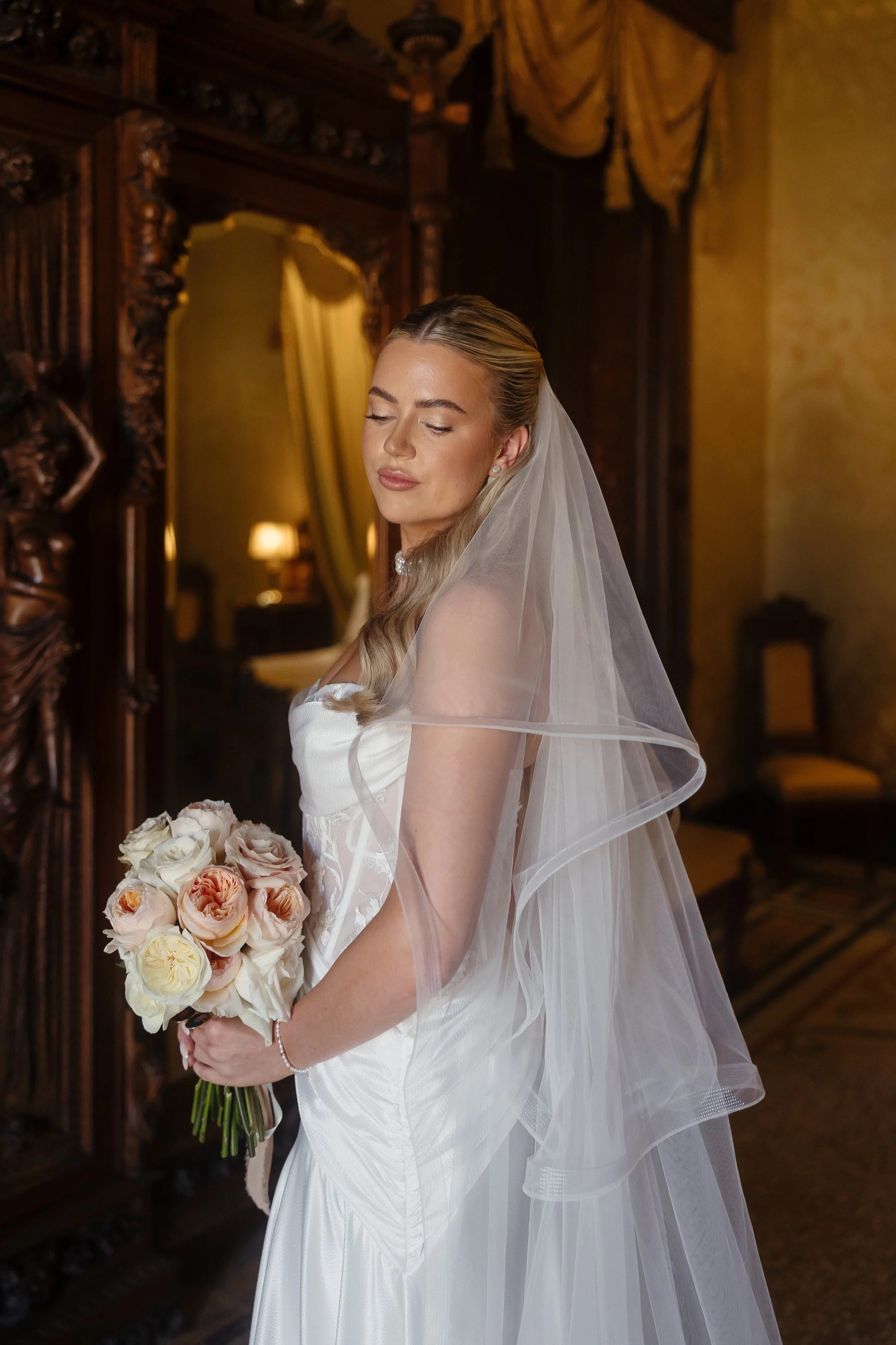 A bride in a white wedding gown holding a bouquet of white and peach roses, standing in a dimly lit vintage room with ornate wooden furniture.