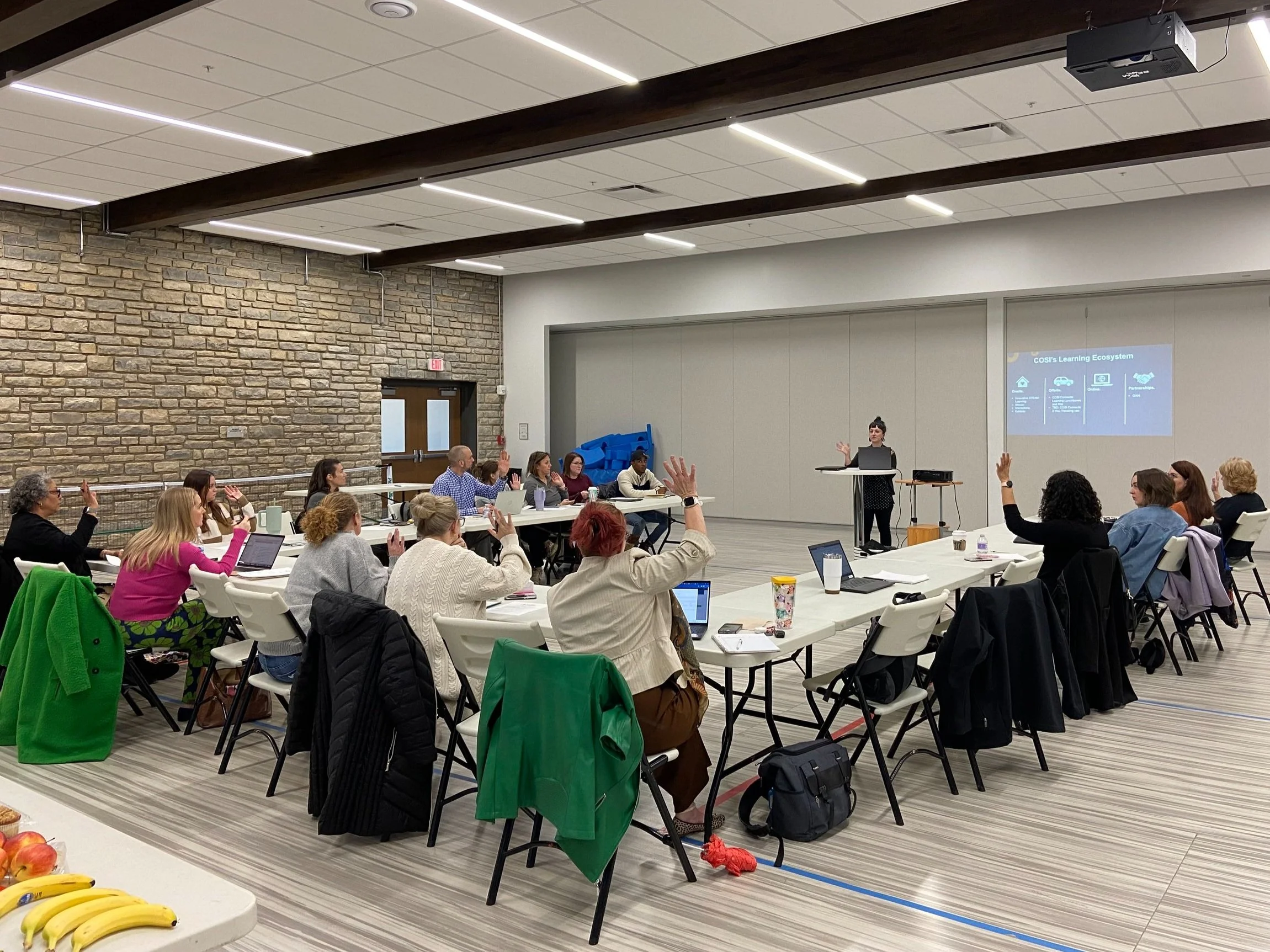 A group of people seated at tables in a meeting room, participating in a presentation. Some have their hands raised. A presenter stands in front of a screen displaying a slide labeled "CDO's Learning Ecosystem." The room has a stone wall and ceiling beams, with snacks on a table in the foreground.