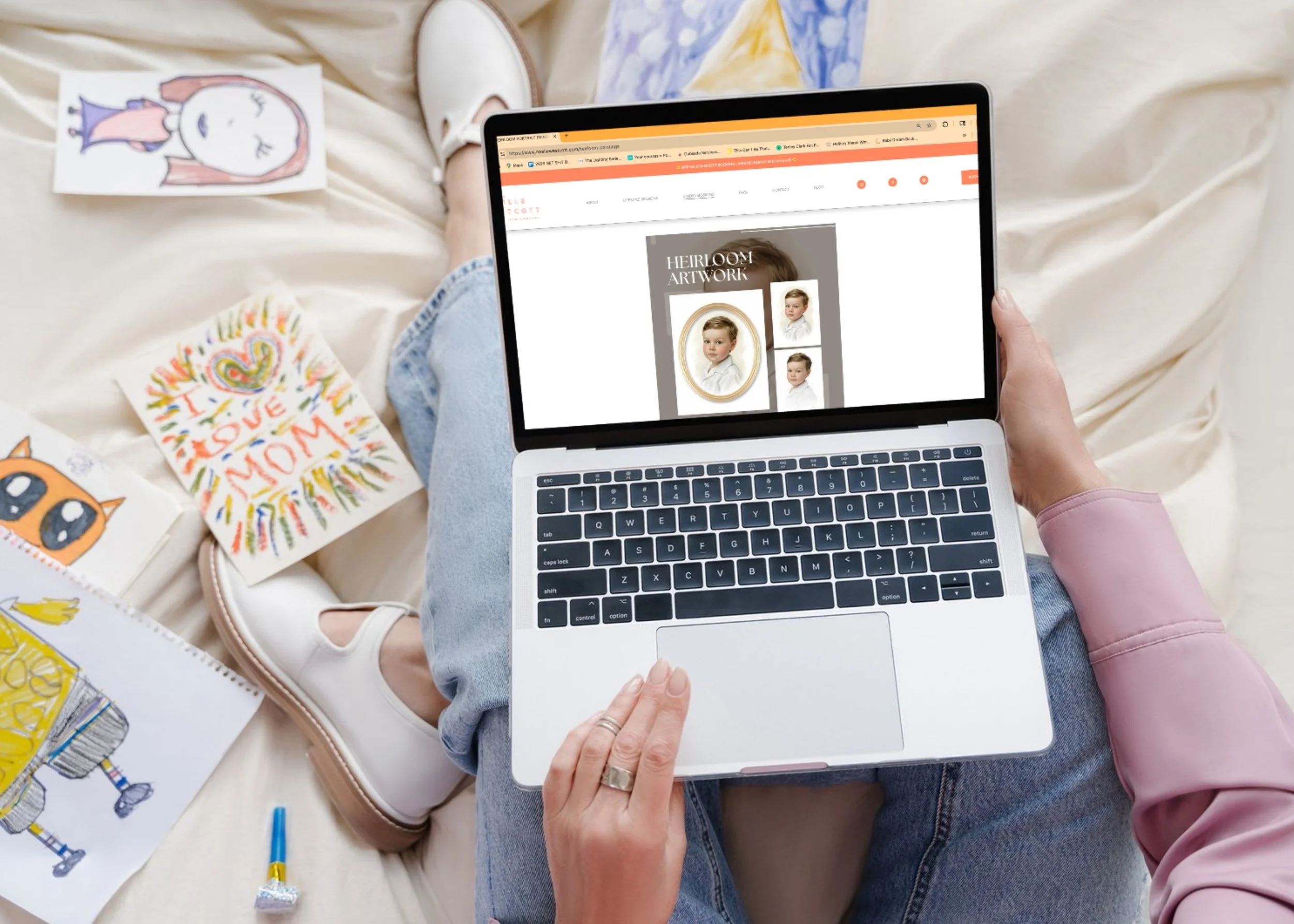 Person sitting on bed using a laptop with a website displaying children's portrait artwork. Around are drawings, a yellow crayon, and a small orange and black cartoon cat drawing.