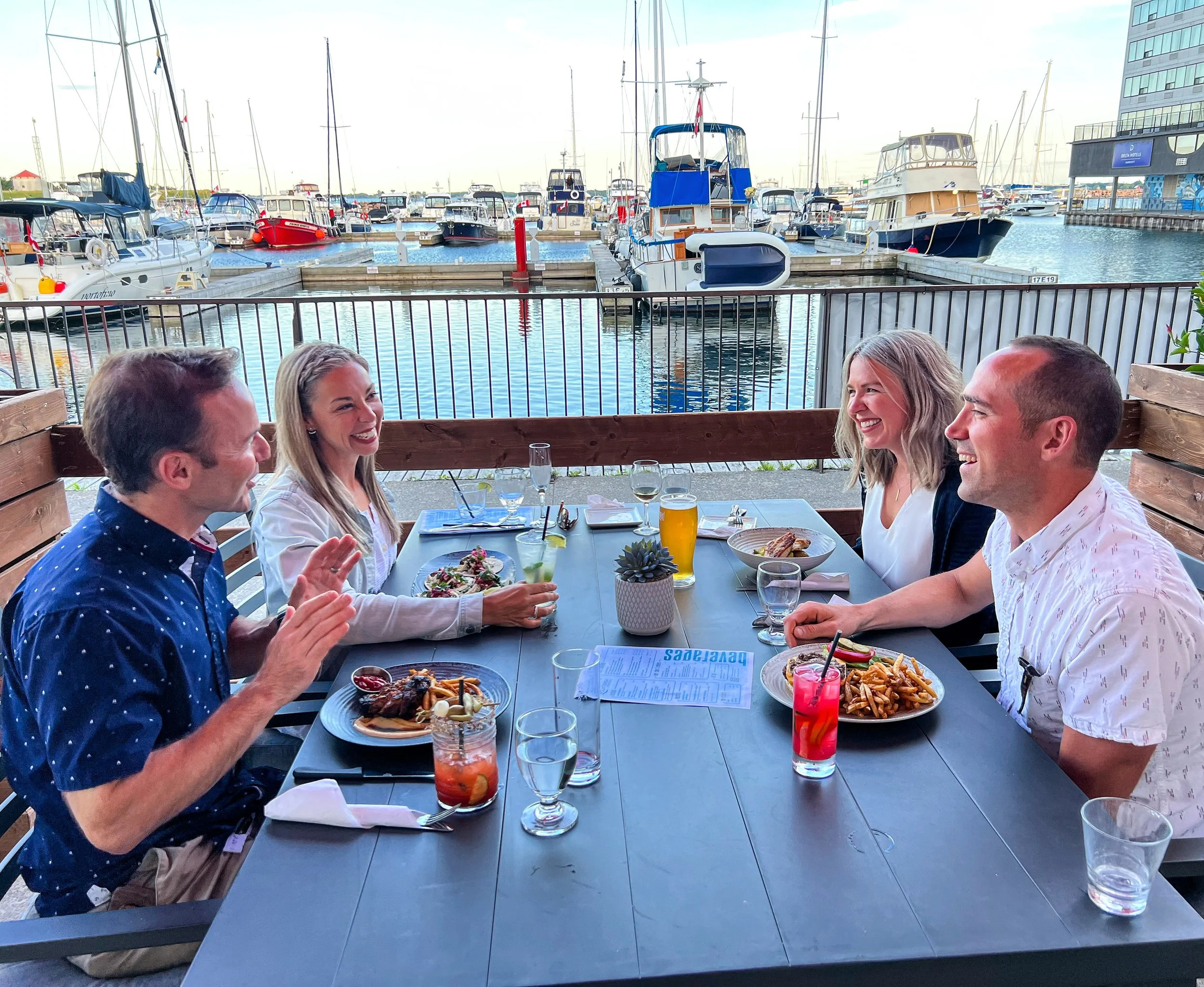Four people enjoying a meal and drinks at an outdoor restaurant with a view of a marina filled with boats.