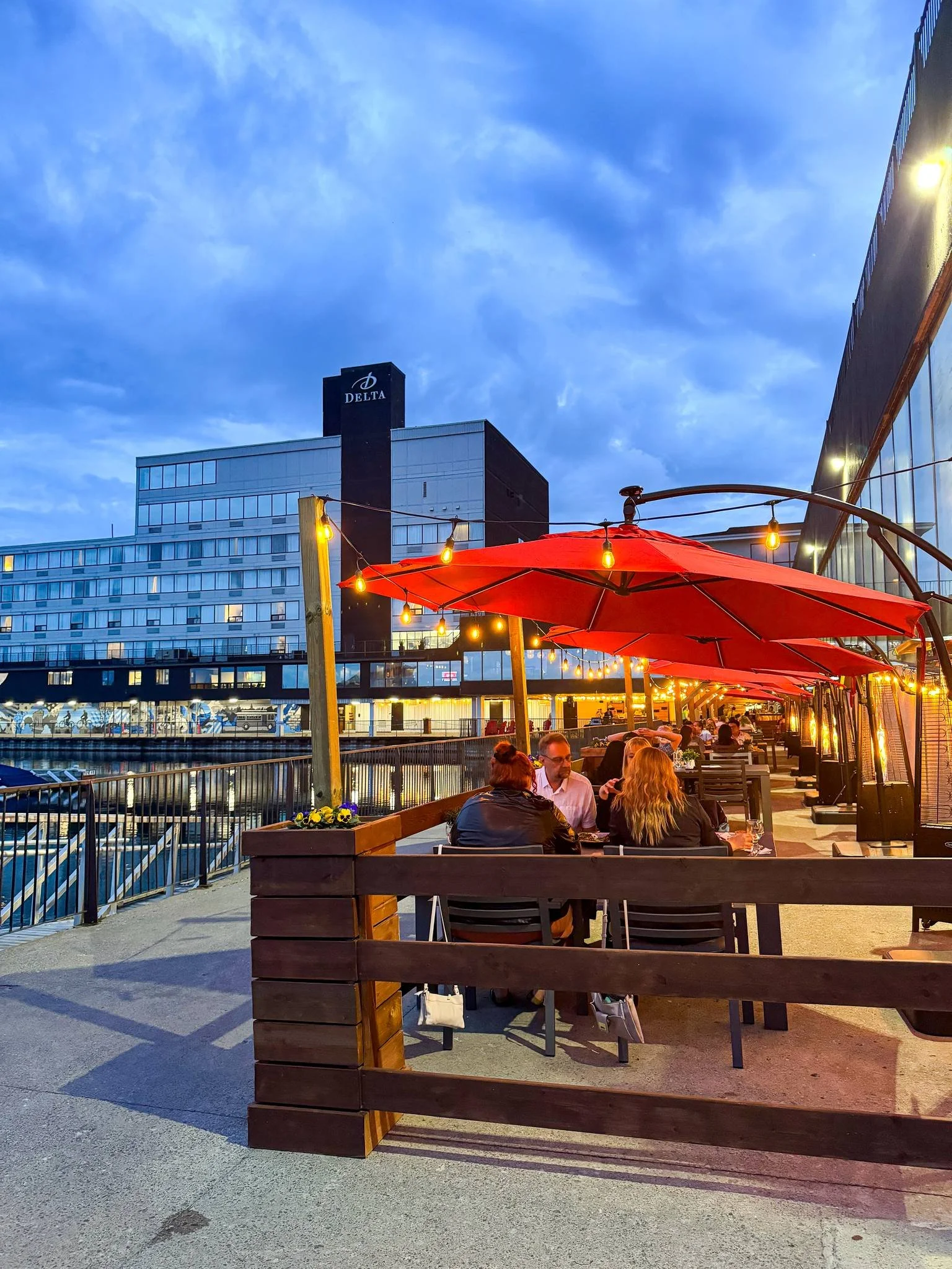 People dining at an outdoor waterfront restaurant with red umbrellas and string lights, overlooking a marina with boats and a modern building with a Delta sign in the background during evening.