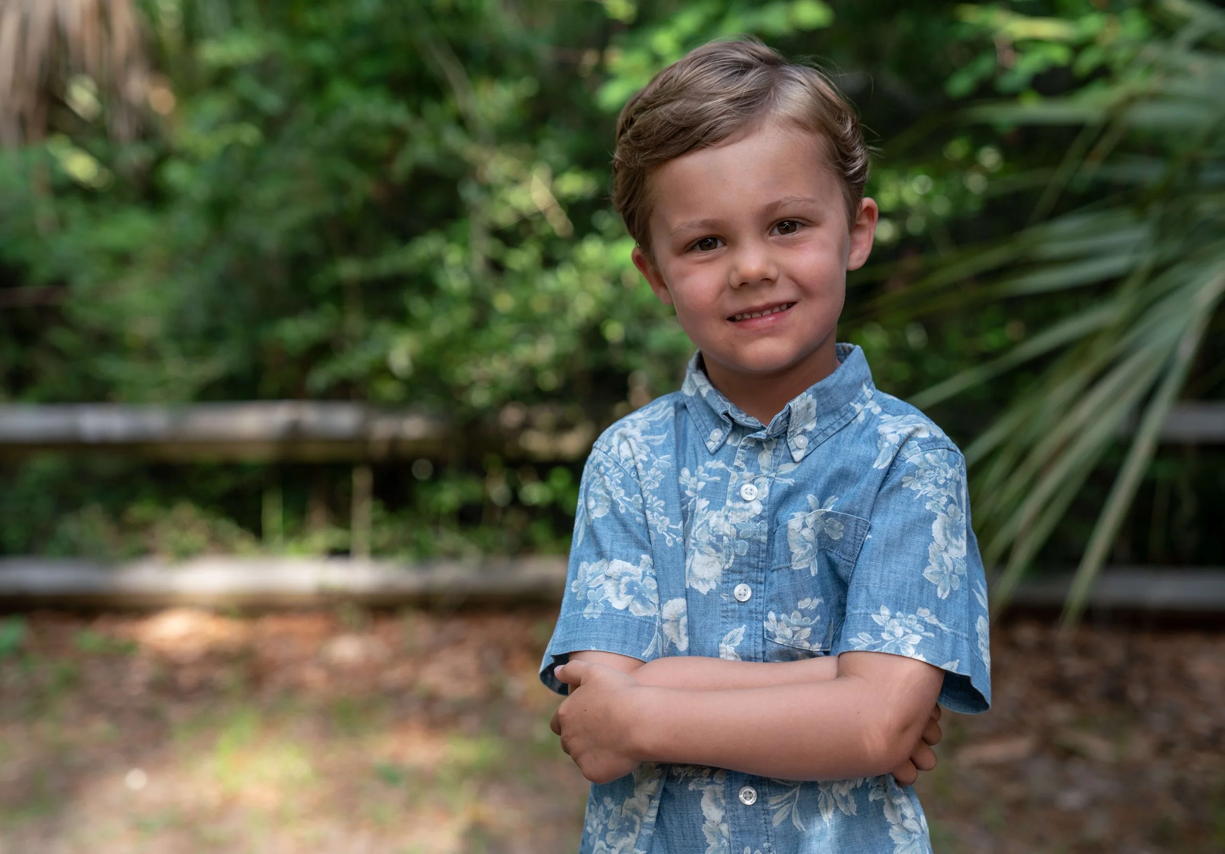 Young boy in a blue shirt standing in front of a fence has his arms crossed and a smile on his face