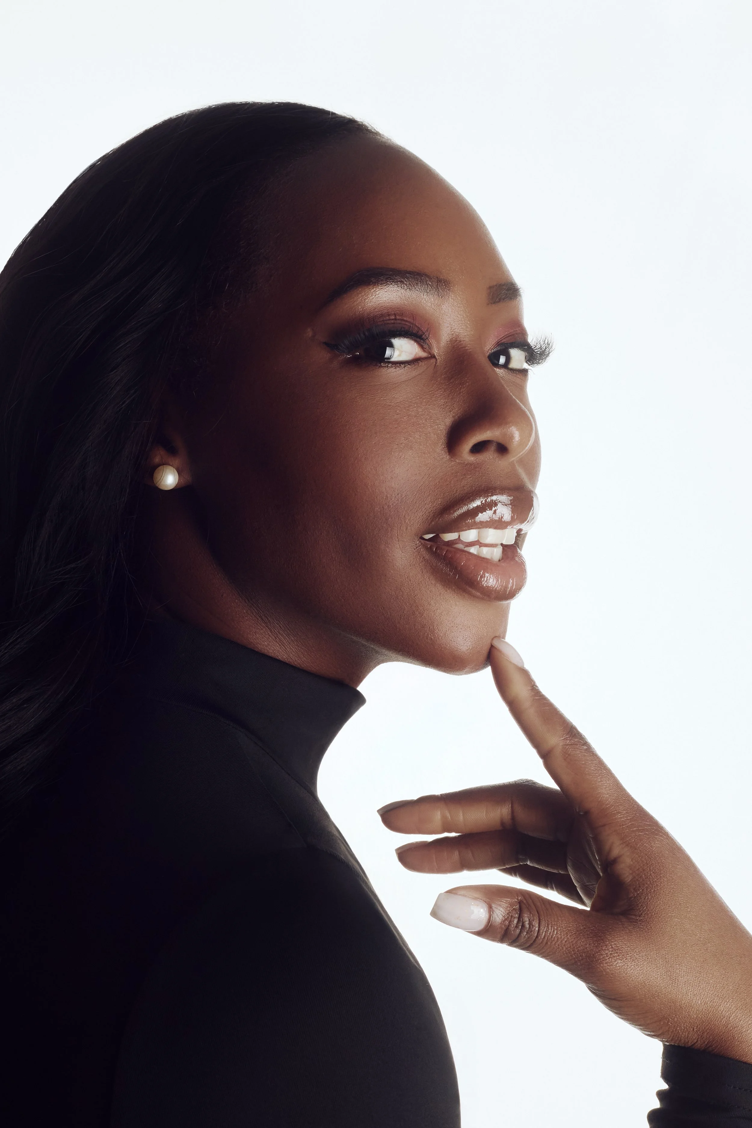 close-up beauty portrait of black woman on photography studio, wearing black turtle neck, long eyelashes, beauty makeup, big lips with shinny lipgloss, white teeth, high-key light, against white background, pearls earrings, Nelson Stein Photography