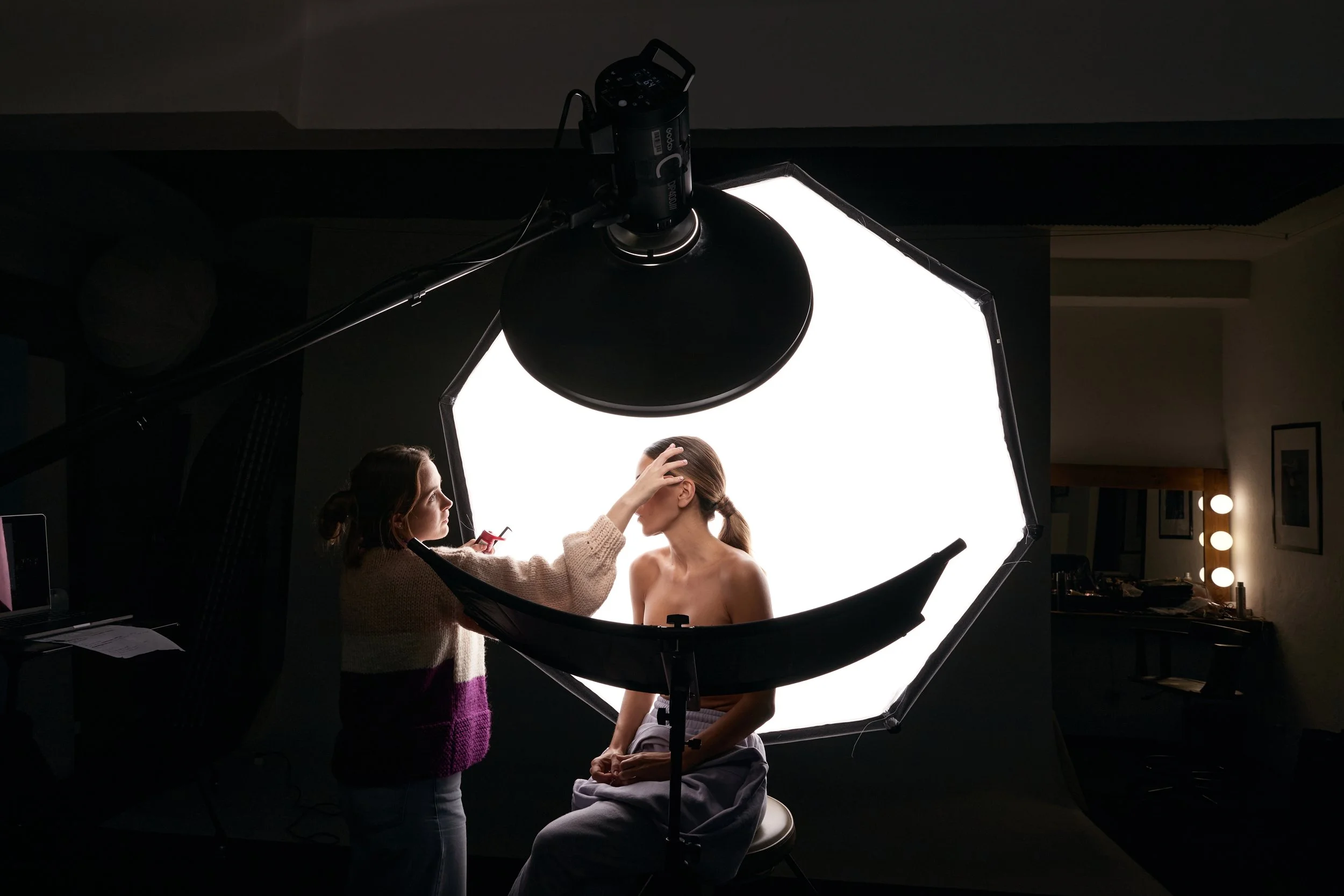 Behind the scenes photo of photography studio lighting set, beauty dish, big octobox, reflector, makeup artist doing the makeup, model on profile, slick hair back, skin showing, white background, Nelson Stein Photography, Lisbon Portugal