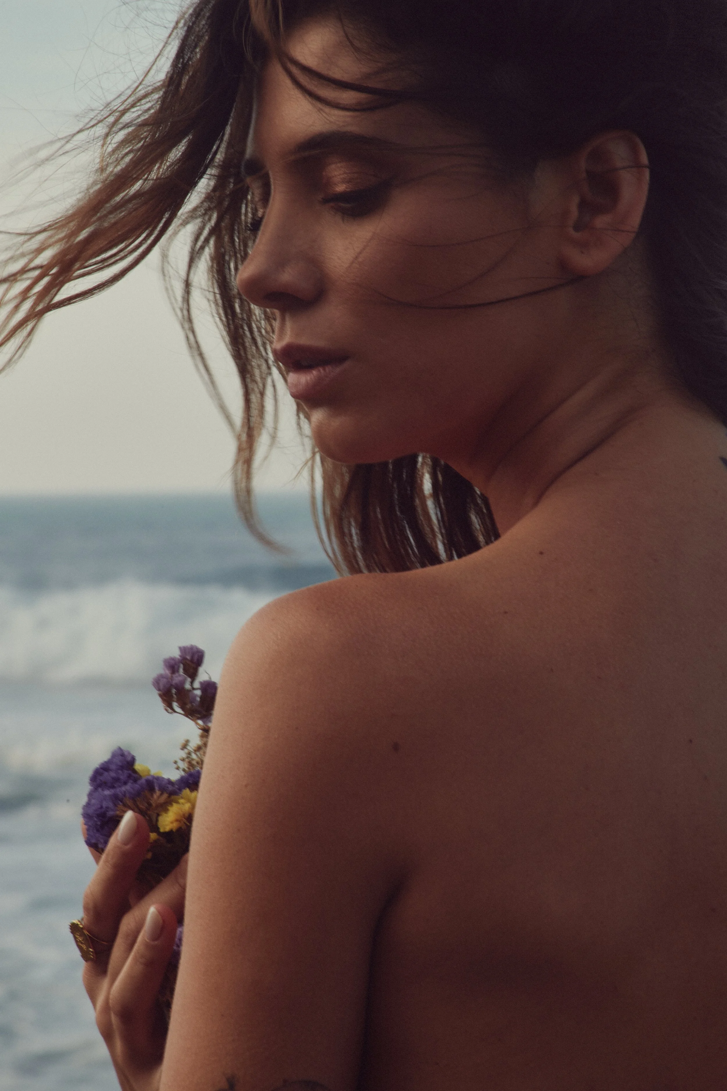 portrait of beautiful brunette model, tanned skin, on beach with ocean behind her, grabbing flowers, delicate facial expression, hair blowing in the wind, model's back to the camera and face turned to camera, Nelson Stein Photography, Lisbon Portugal