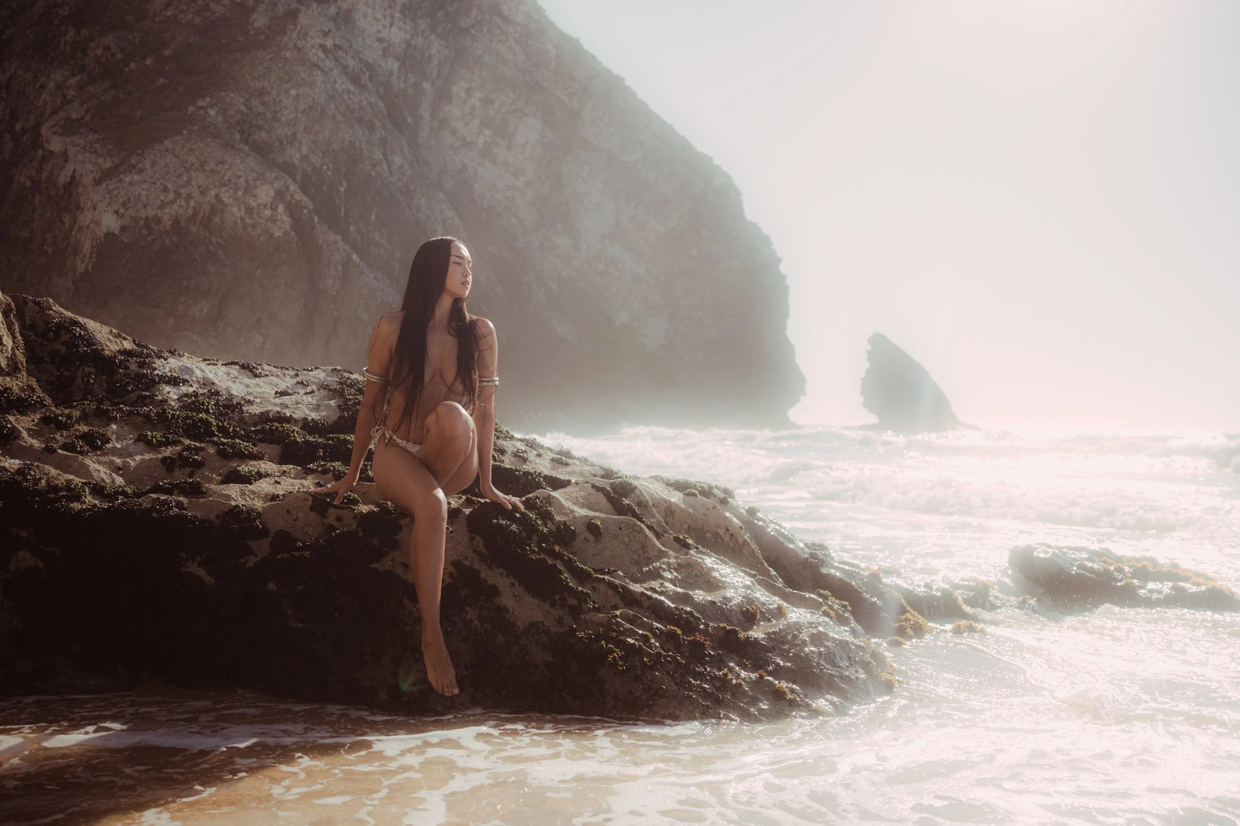 portrait of a beautiful woman with dark long hair sat on a rock at the beach, nude, background of big rocks and sea water, mermaid, natural light, magical color grading, Nelson Stein Photography, Lisbon Portugal