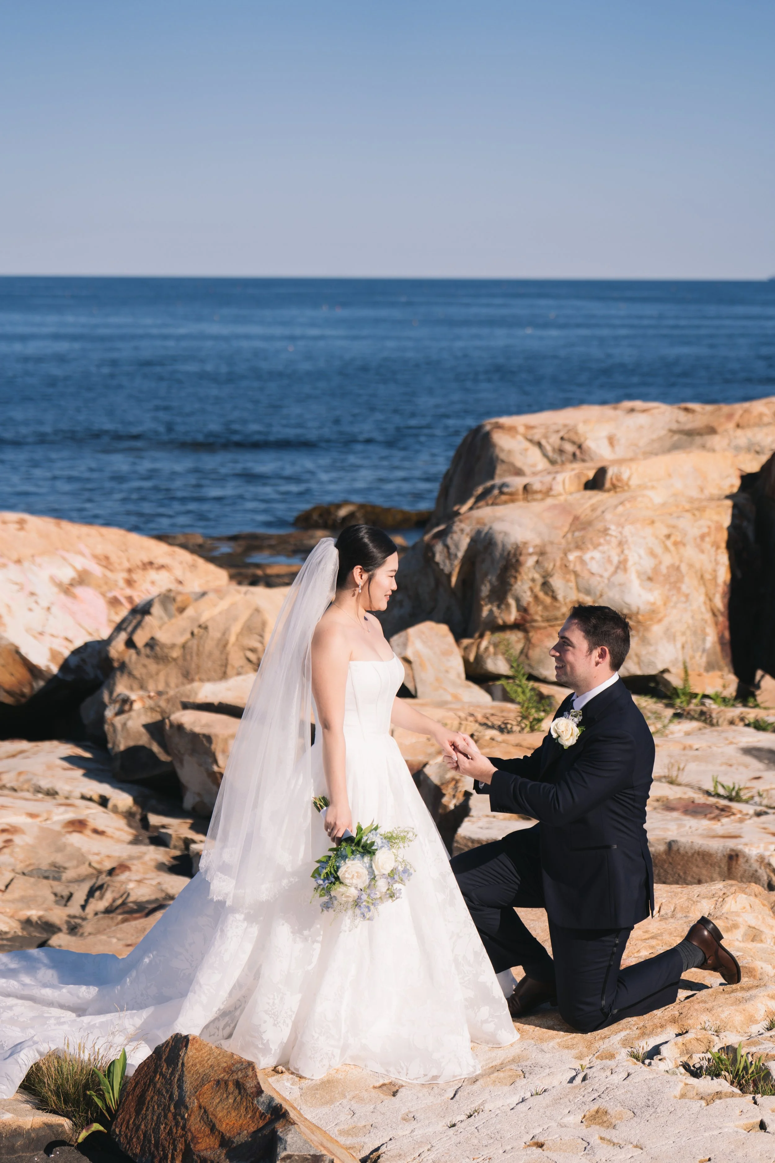 A bride and groom on a rocky beach, with the groom kneeling on one knee, holding the bride's hand, during their wedding vows with the ocean in the background.