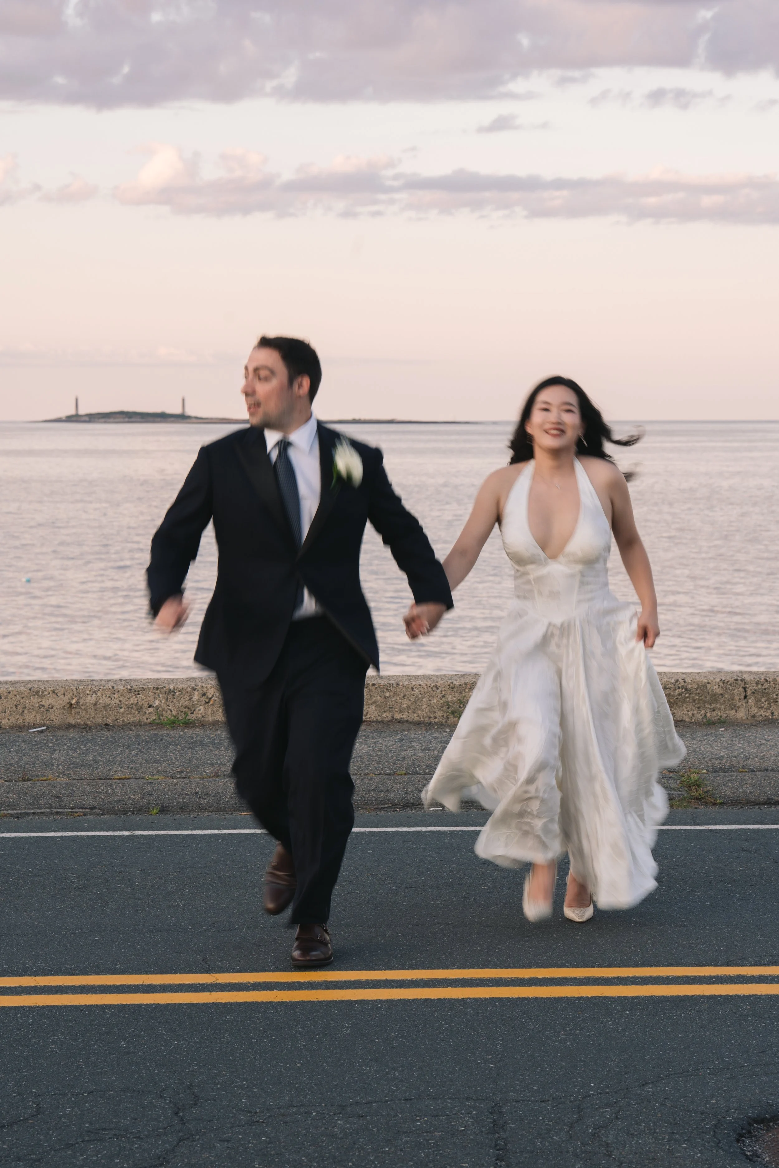 A happy couple, dressed in wedding attire, running hand in hand along a waterfront promenade at sunset, with the ocean and cloudy sky in the background, capturing a joyful moment.