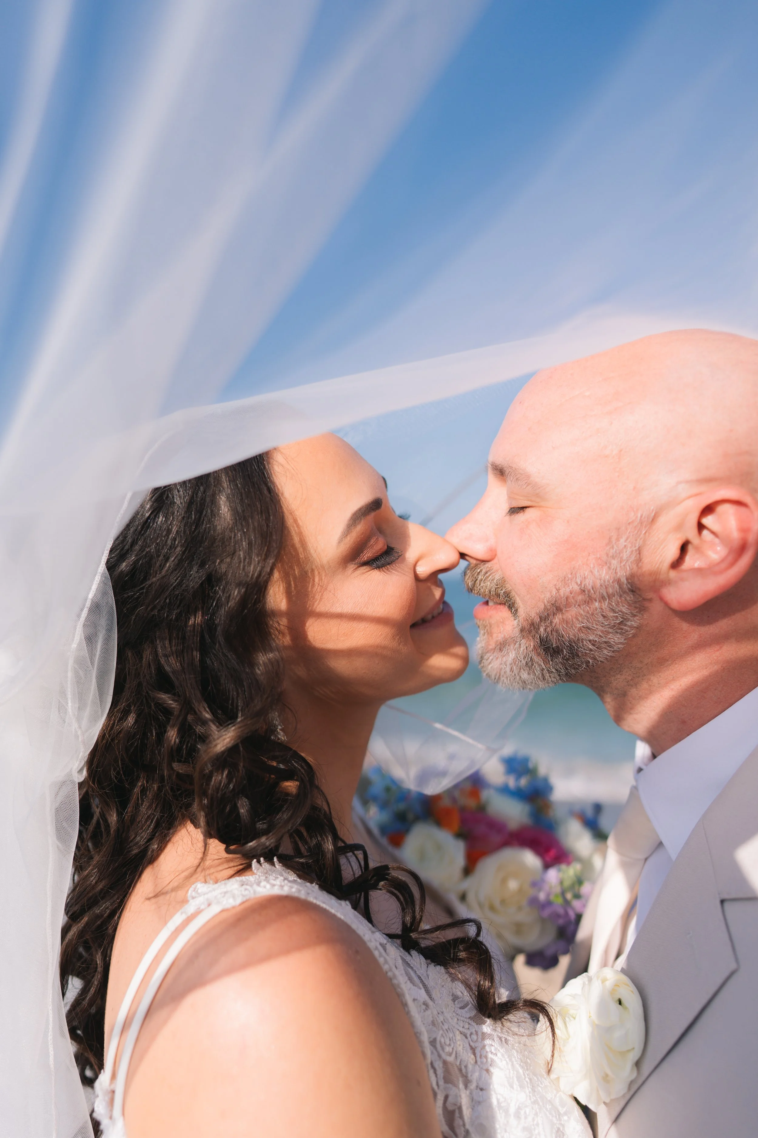 A bride and groom facing each other with noses touching, outdoors on a sunny day, with the bride wearing a veil and lace dress, and the groom in a light-colored suit with a boutonniere of white flowers.