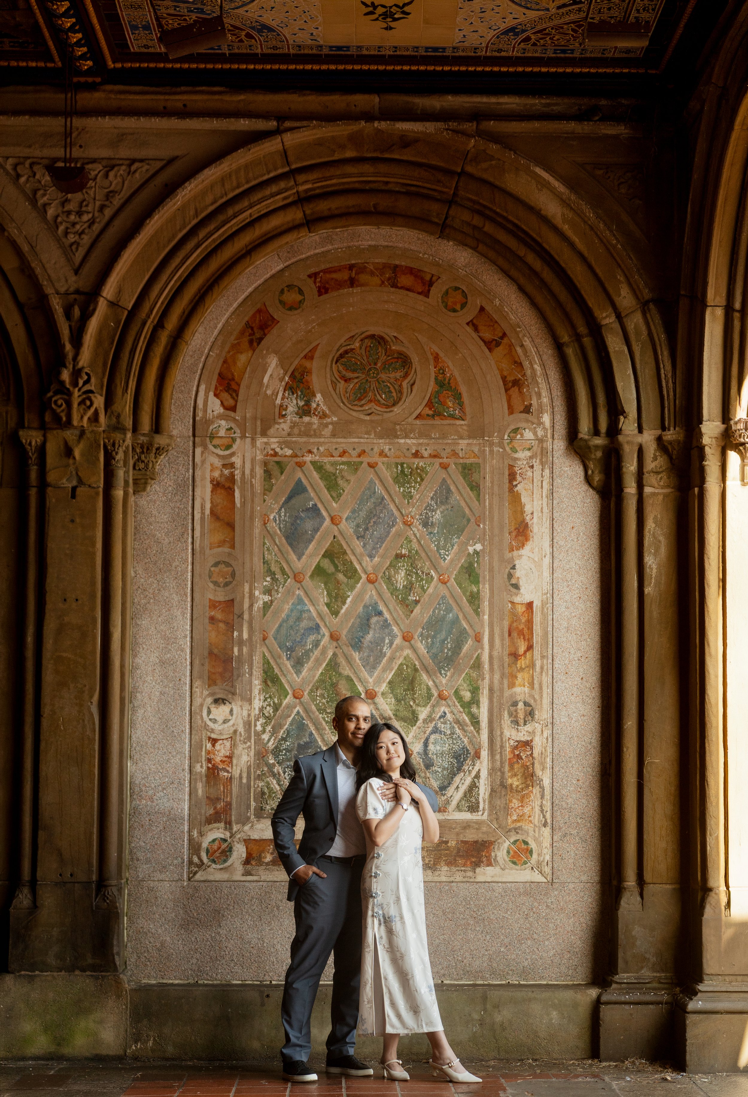 A man and woman pose together in front of a decorative old building with ornate archways and intricate woodwork. Their engagement photos at the Bethesda Terrace in Central Park of New York City.