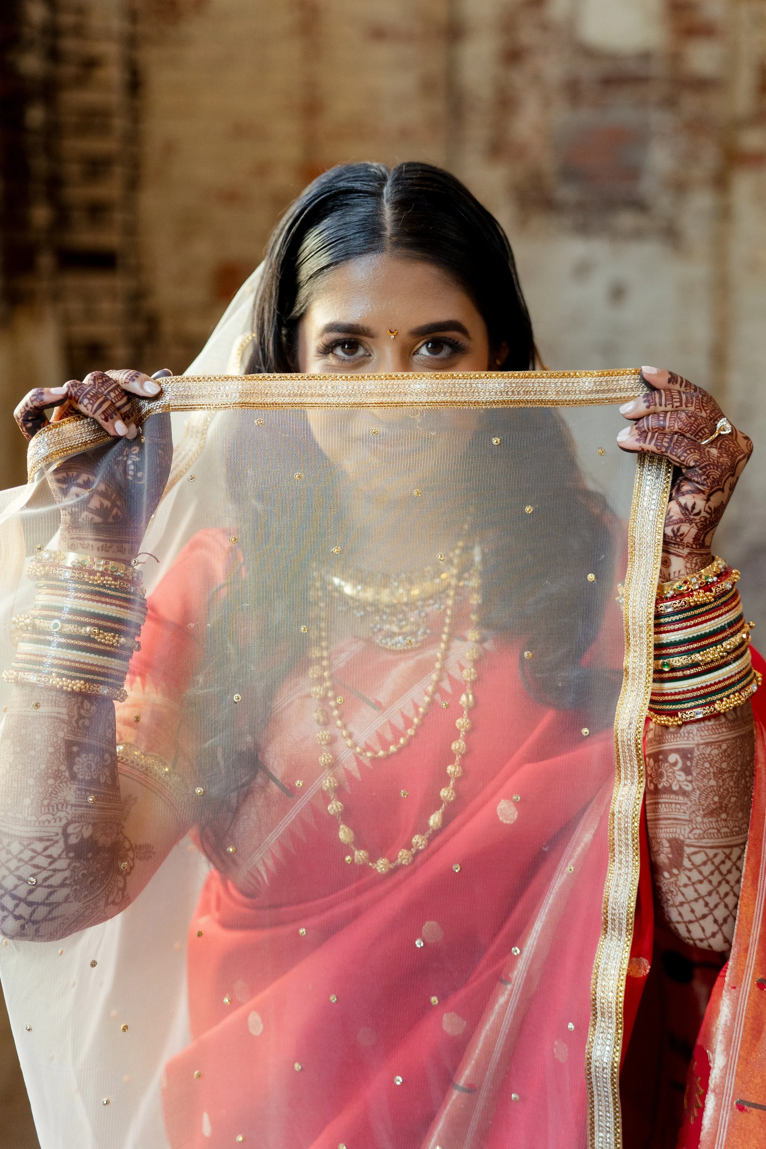 A Indian bride with dark hair and brown skin peering over a sheer, gold-embellished pink and cream dupatta, adorned in traditional Indian jewelry and clothing, standing against a rustic brick wall. Wedding at Guardian Works in Atlanta, GA.