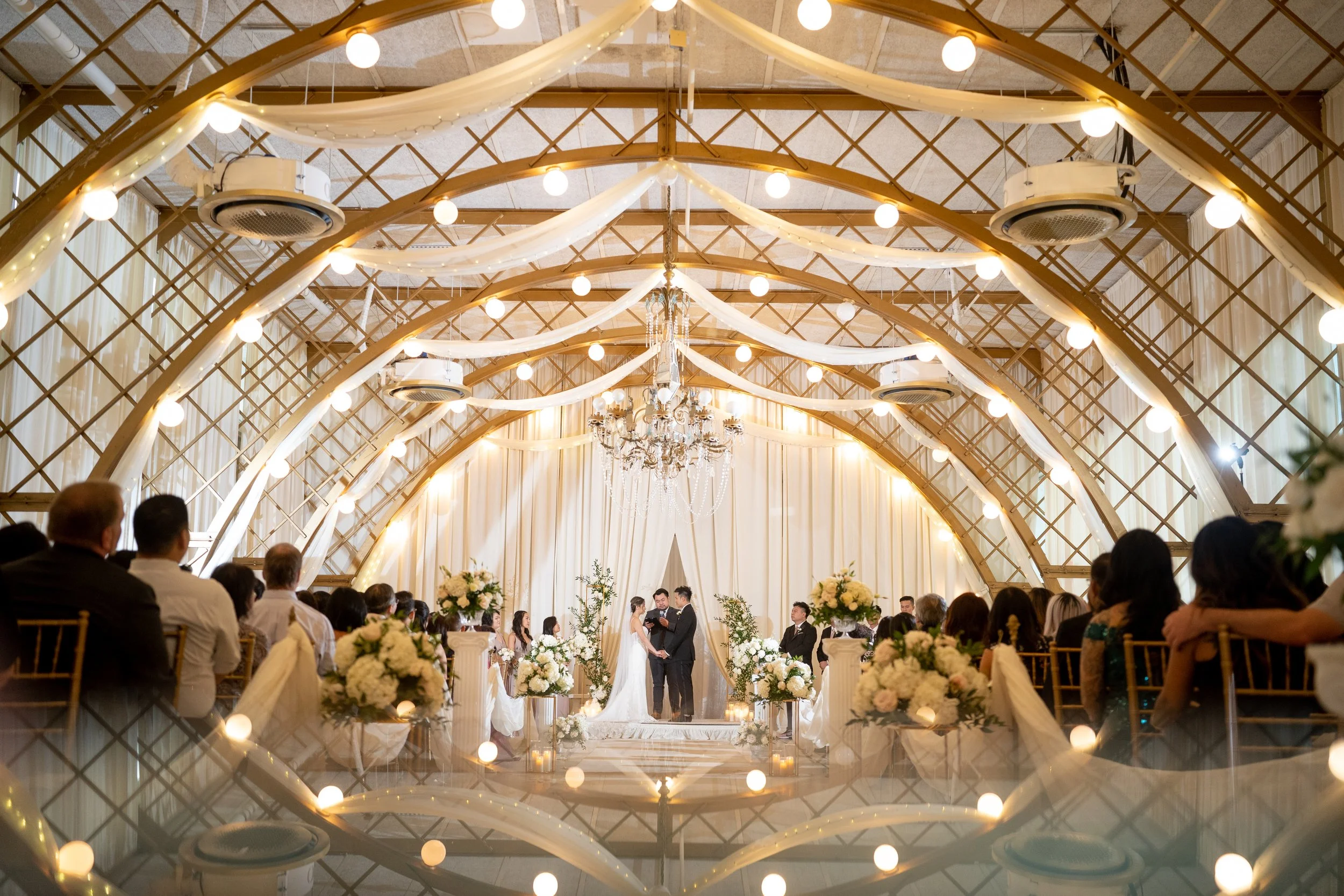 Wedding ceremony with bride and groom exchanging vows in a decorated indoor venue, with guests seated on both sides, floral arrangements, draped fabric, and a chandelier, viewed through a decorative mirror or reflective surface.