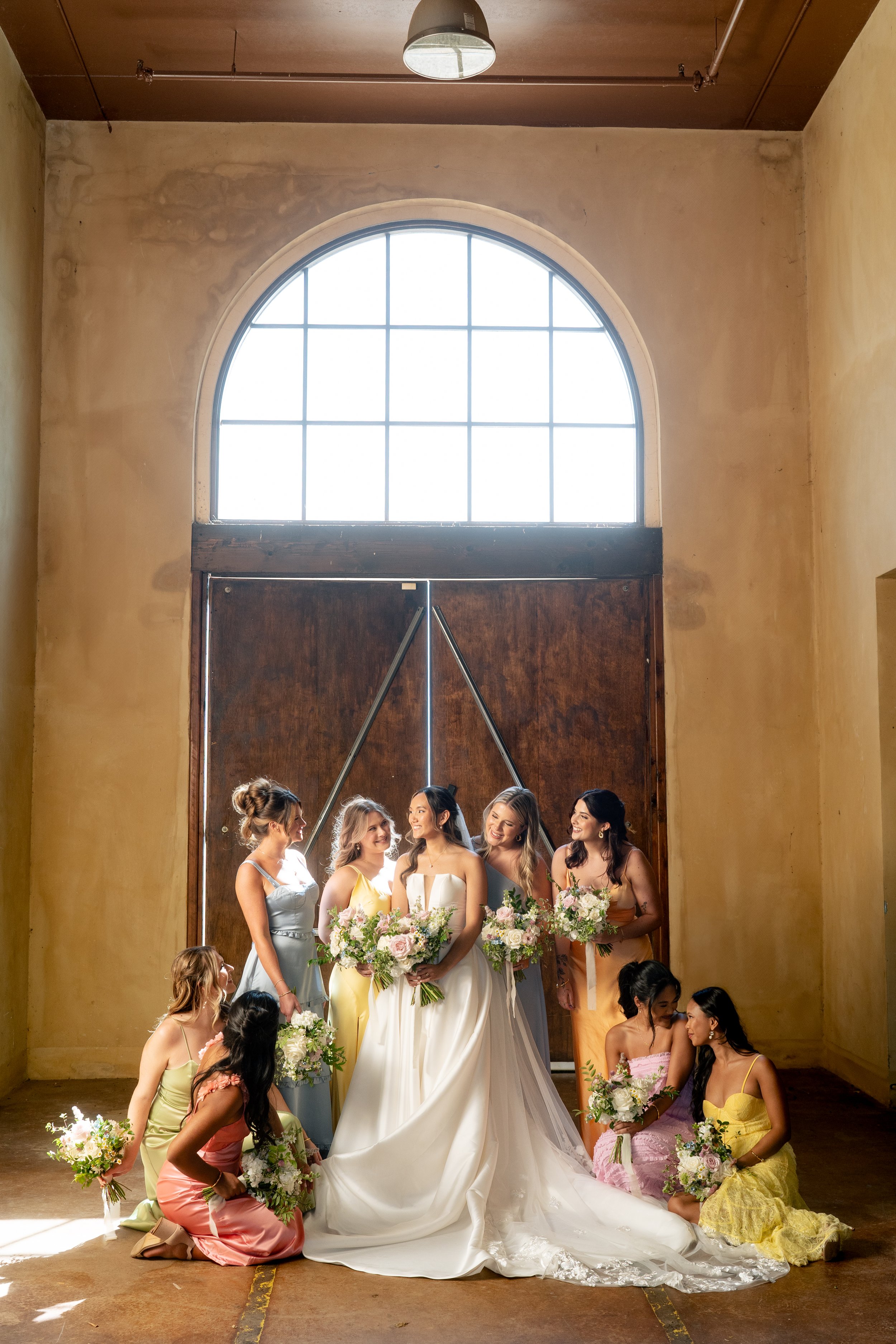 A bride and her bridesmaids in colorful dresses in a spacious room with large arched window and wooden door at Montaluce Winery, holding bouquets of flowers.