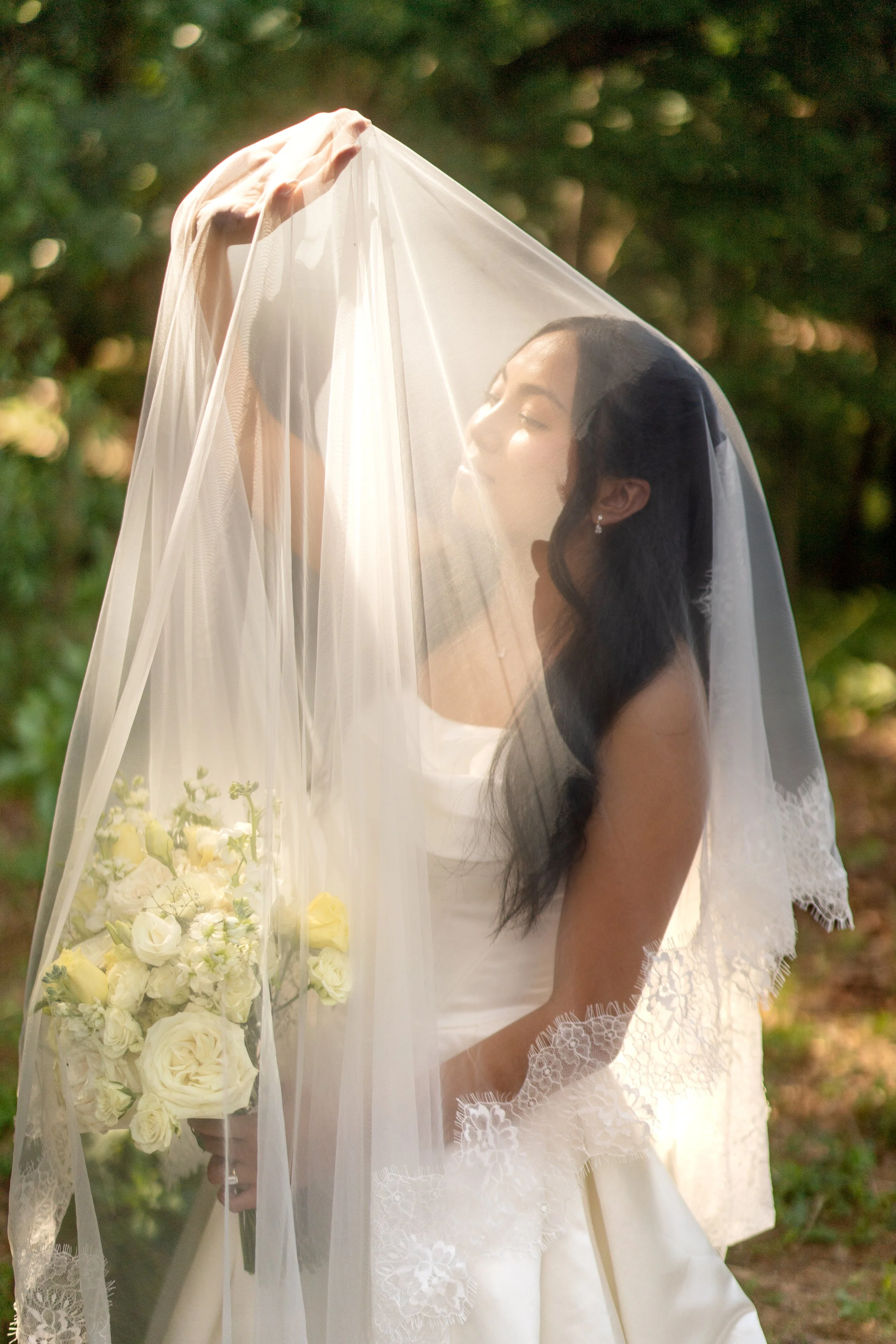 A bride with dark hair and earrings holding a bouquet of white and cream flowers, with veil over her face outdoors at a Venue in Brooklyn NYC. Photo by Aurora Photography a NYC wedding photographer.