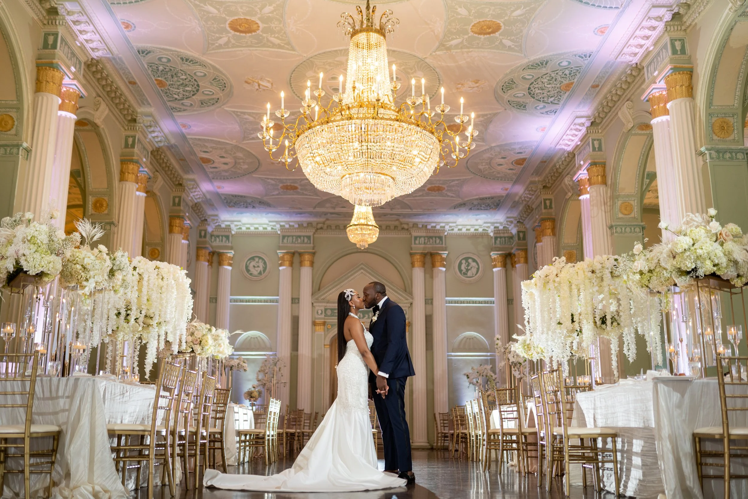 A bride and groom in wedding attire sharing a kiss in an elegant decorated wedding hall at the Biltmore Ballrooms with a large chandelier, floral arrangements, and gold chairs.
