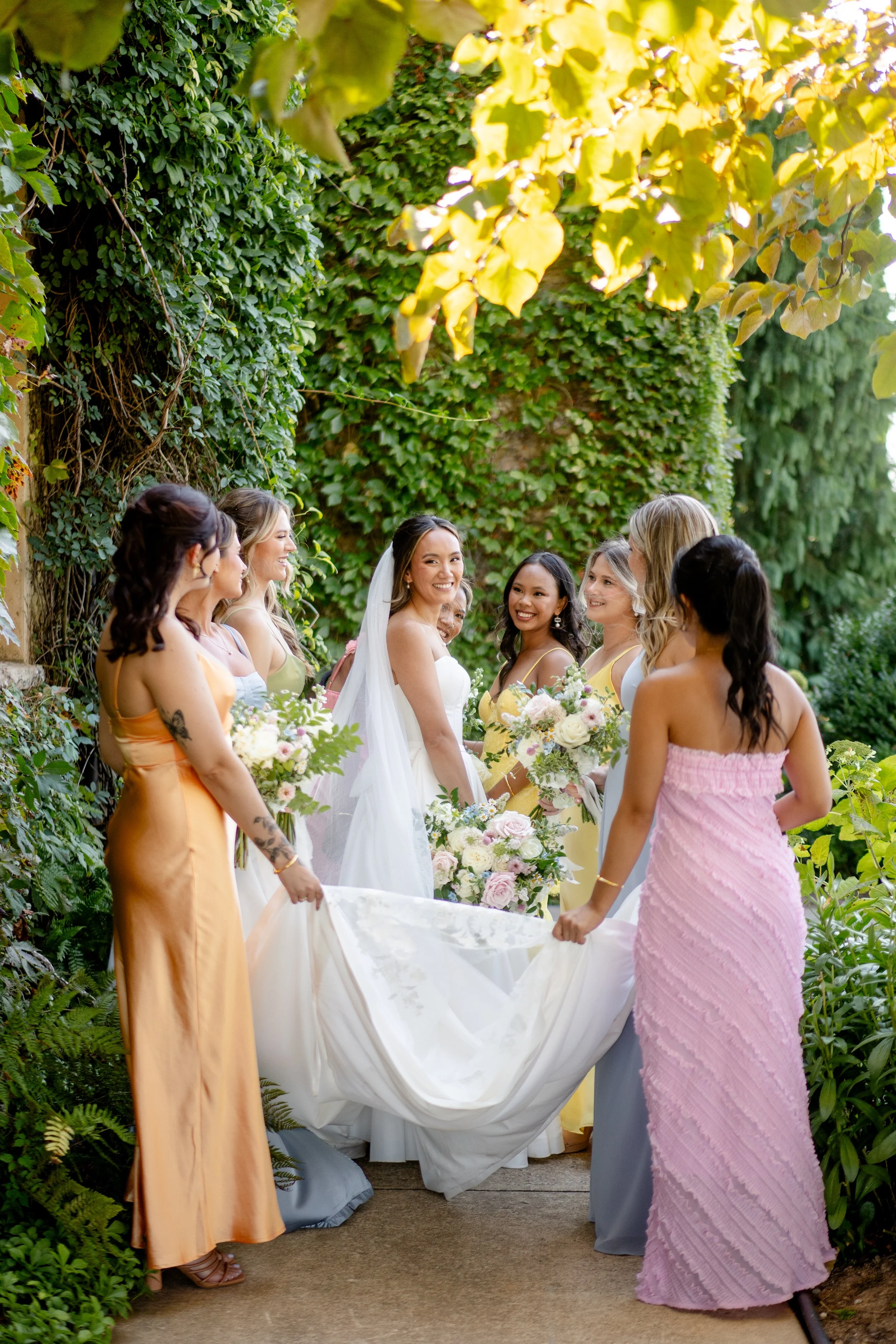A bride at Montaluce Vineyards in a white wedding dress with a veil and bouquet standing in the center, surrounded by bridesmaids in colorful dresses, holding flowers, outdoors in front of a lush green ivy-covered wall, celebrating a wedding.