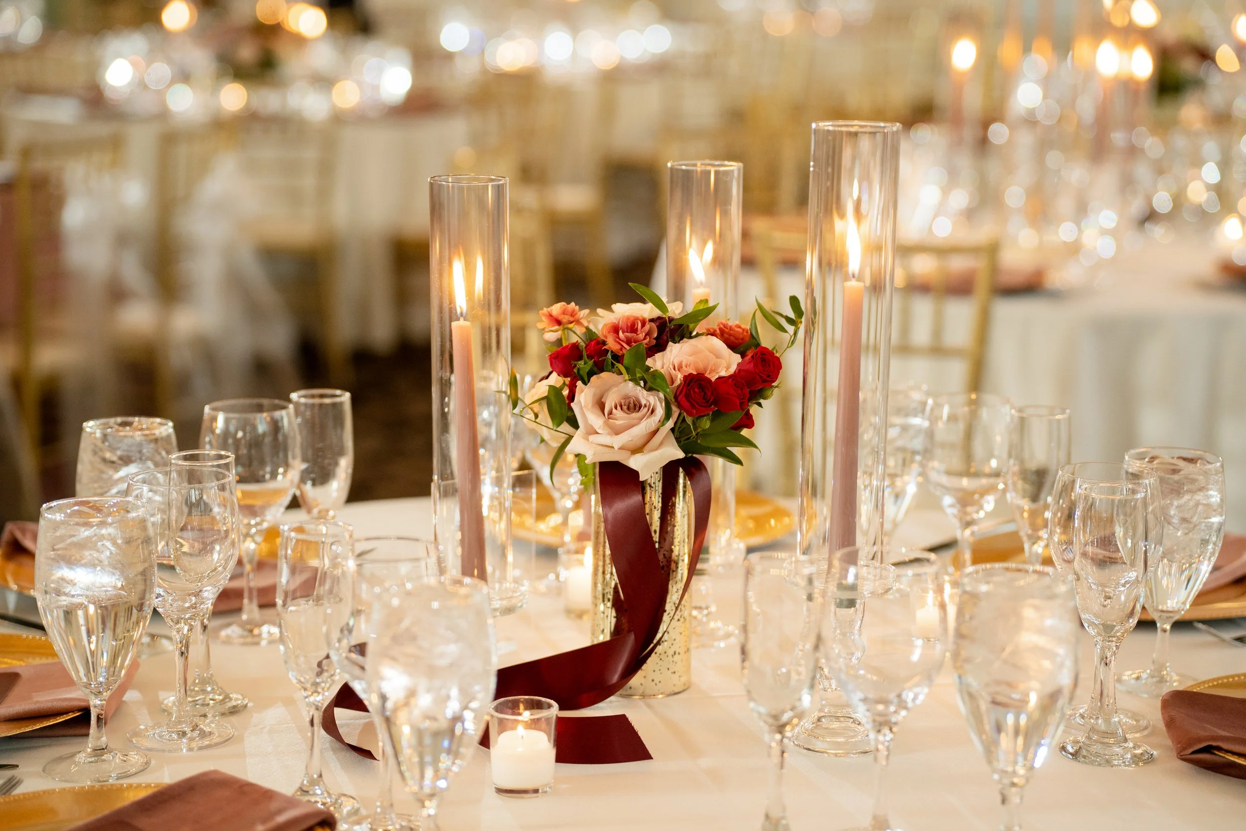 Elegant banquet table decorated with pink and red flowers in a gold vase, surrounded by multiple wine glasses and candles, set in a warmly lit event hall.