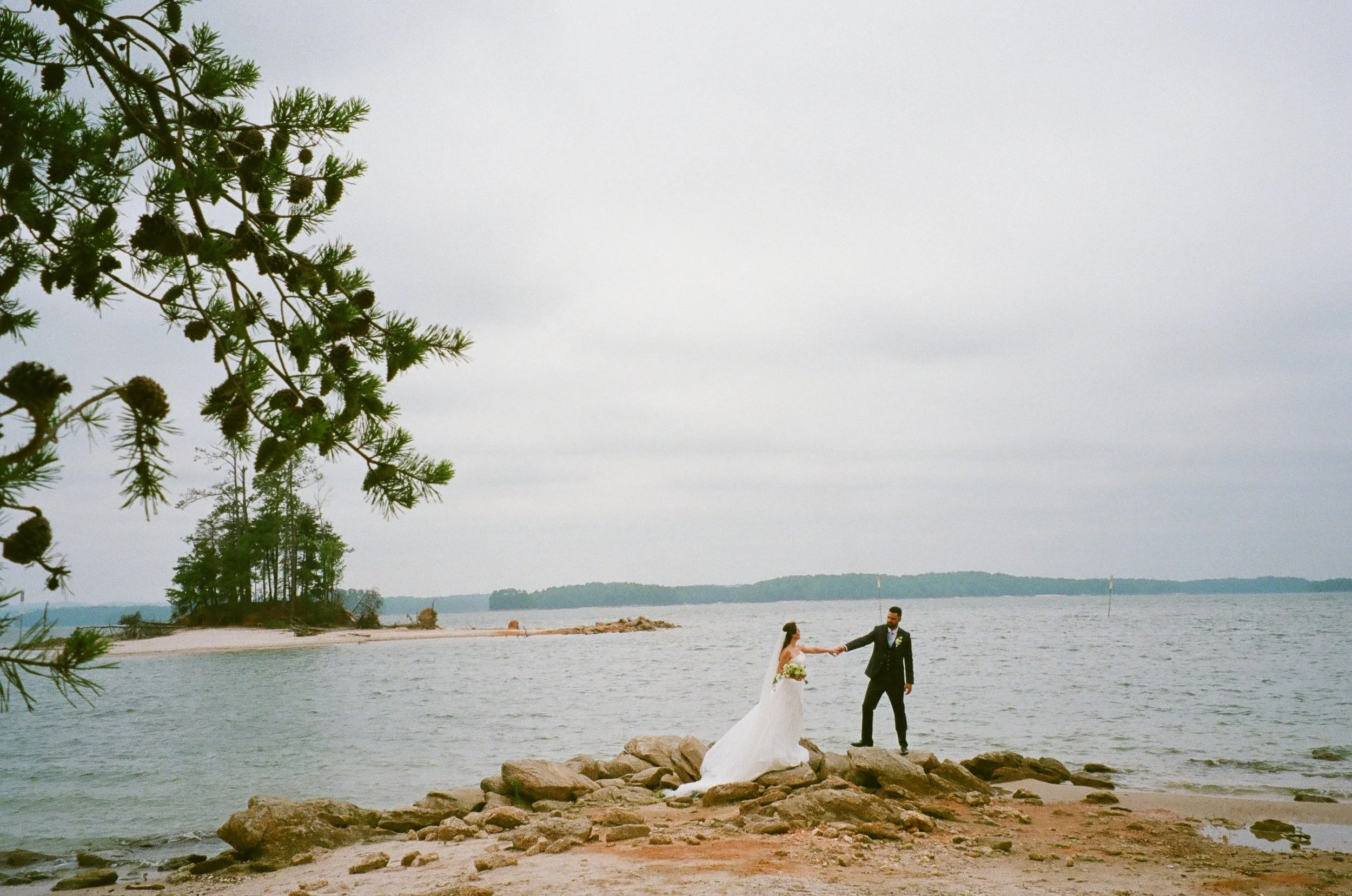 A film photo by Aurora Photography of a  bride and groom holding hands on a rocky beach at Lake Lanier Island, with water and distant land in the background, during their wedding.
