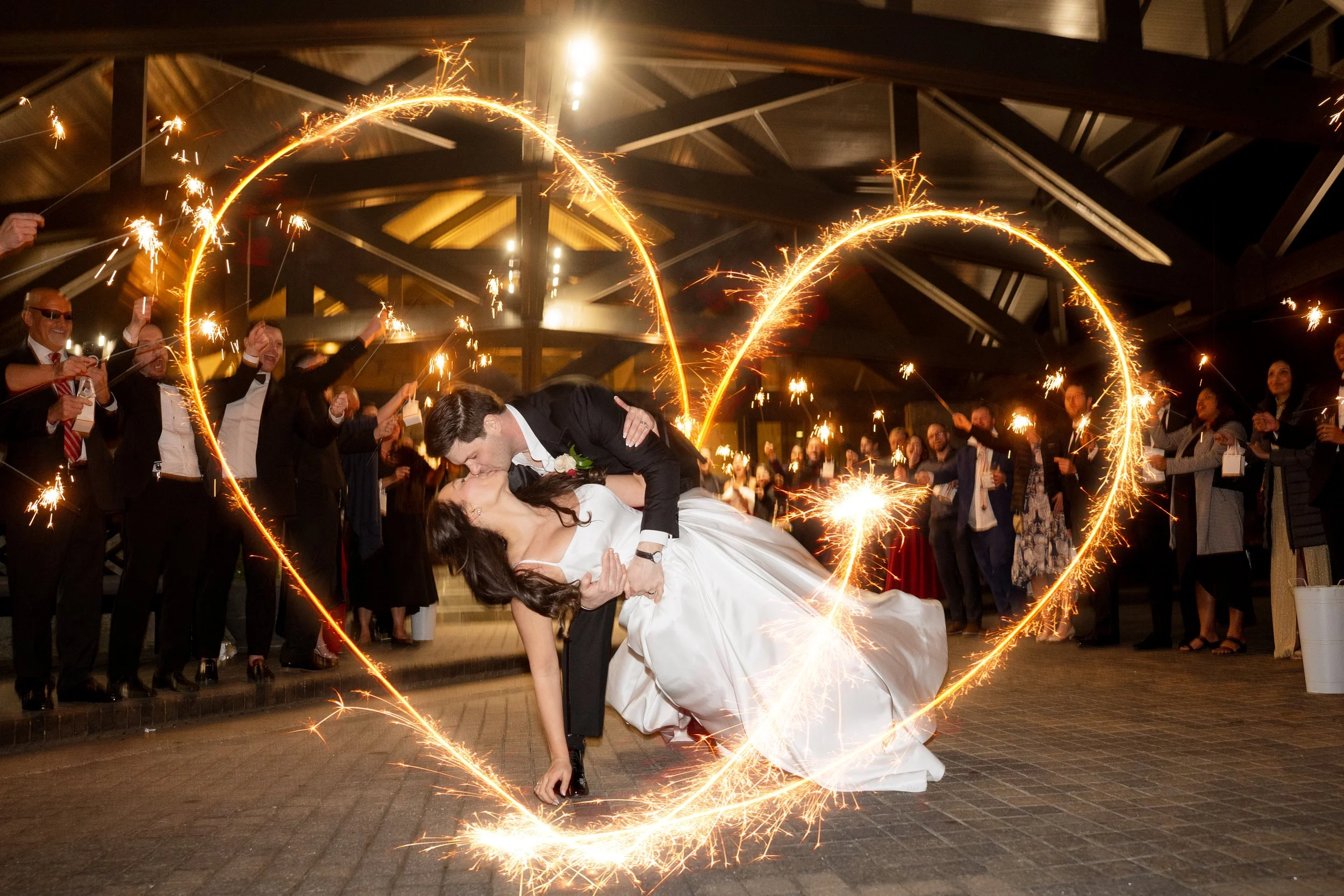 A newlywed couple sharing a kiss during their send off at Country Club of the South, surrounded by friends and family holding sparklers, creating a heart shape with the sparklers in the night.