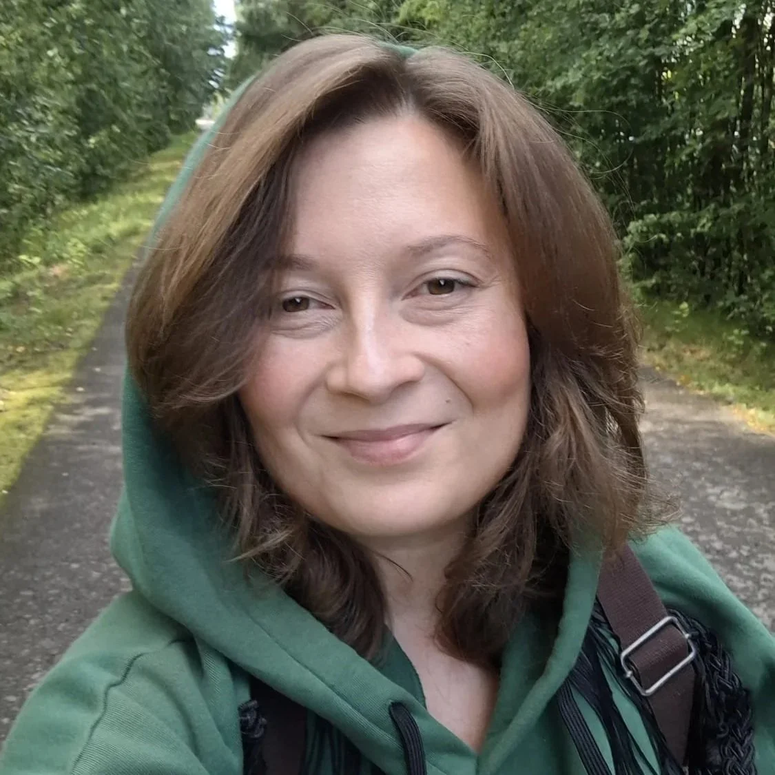 Nainen hymyilee ulkona metsässä kävellessään. Hänellä on ruskeat hiukset ja vihreä huppari.