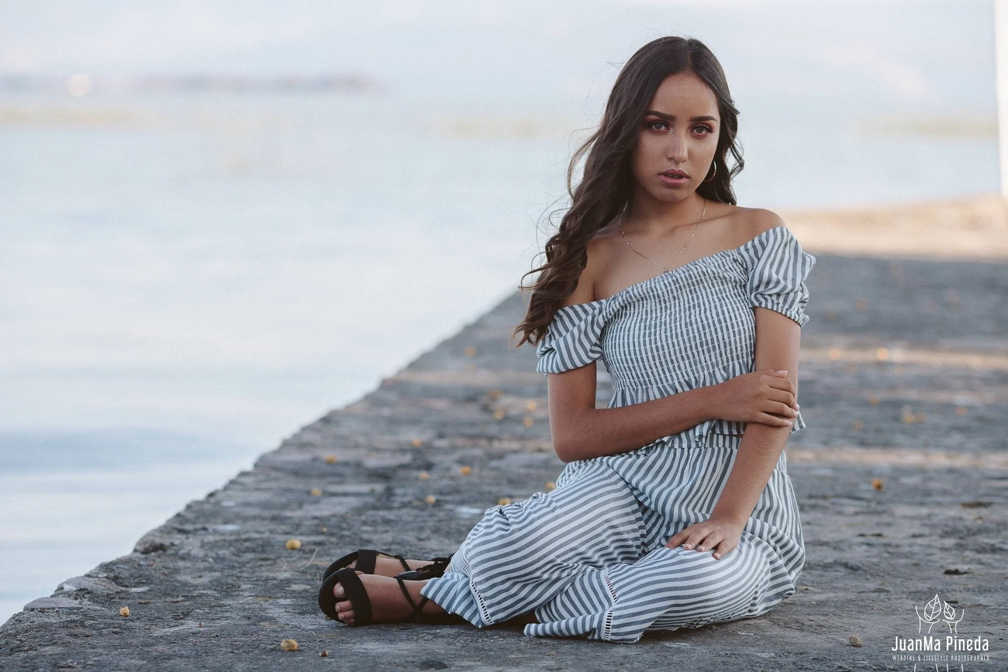 Mujer sentada en un muelle frente al agua, con vestido de rayas y sandalias negras.
