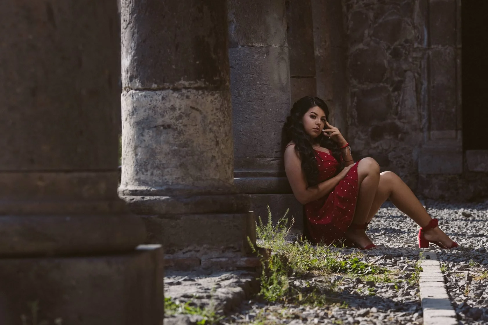 Mujer sentada junto a una columna de piedra, con vestido rojo y zapatos a juego, en un ambiente al aire libre.