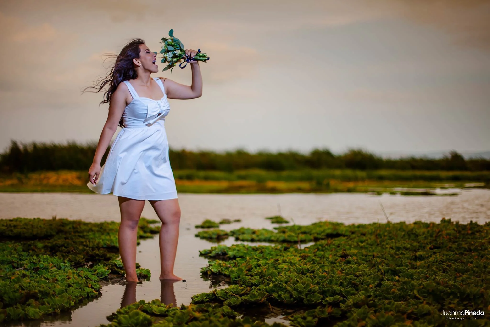 Mujer con vestido blanco sosteniendo un ramo de flores en un paisaje con agua y plantas acuáticas.