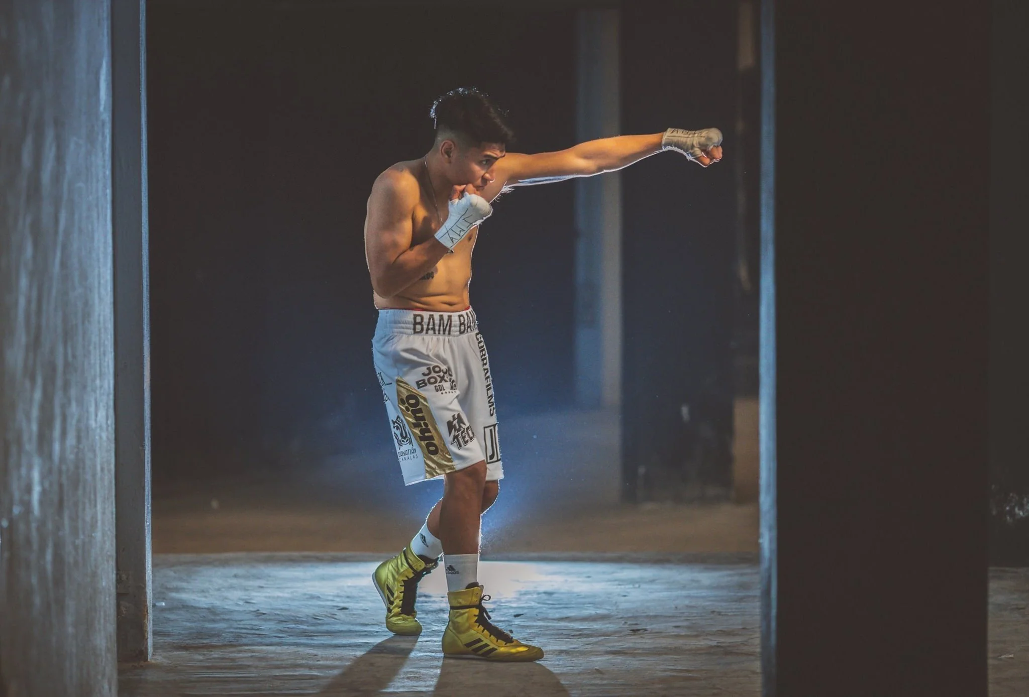 Boxeador entrenando en un gimnasio oscuro, lanzando un puñetazo, vistiendo pantalones cortos blancos y guantes de vendaje, con zapatillas deportivas amarillas.