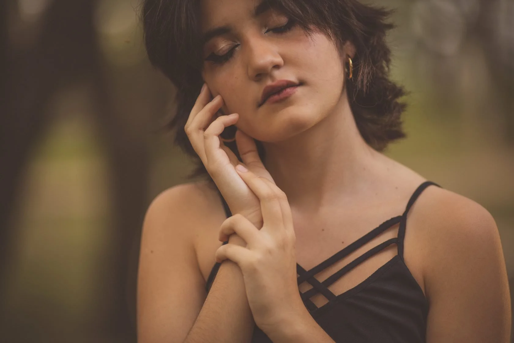 Mujer joven con ojos cerrados y expresión tranquila, tocando su rostro con las manos, en un entorno natural.