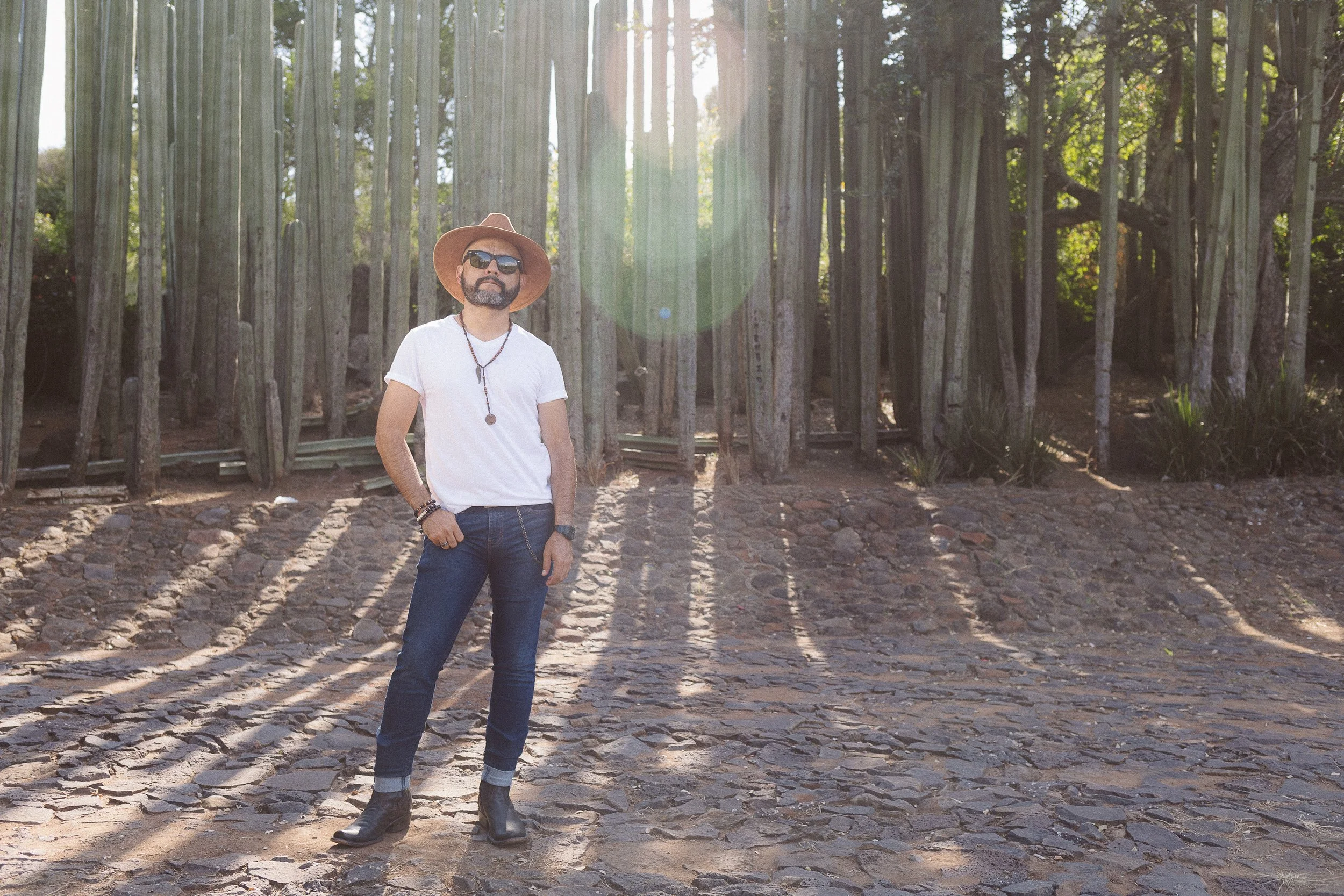 Hombre con sombrero de vaquero, camisa blanca, jeans y botas, de pie en un entorno natural con cactus altos y piedras en el suelo, iluminado por la luz del sol.