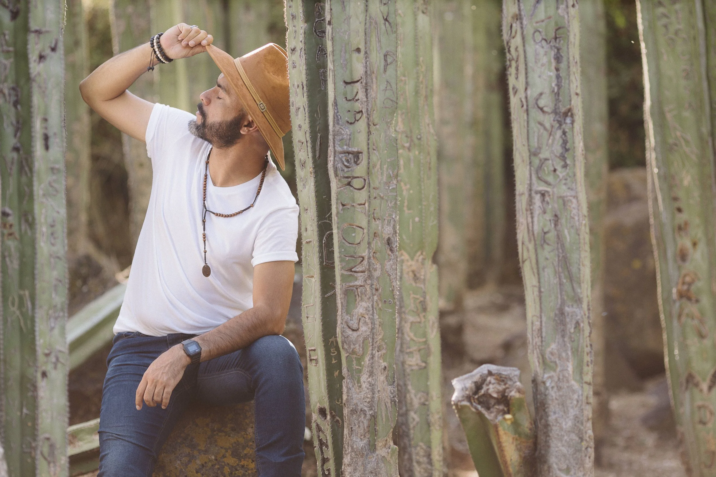 Hombre con sombrero en un jardín de cactus, sentado, luciendo una camiseta blanca y jeans, rodeado de cactus grabados.